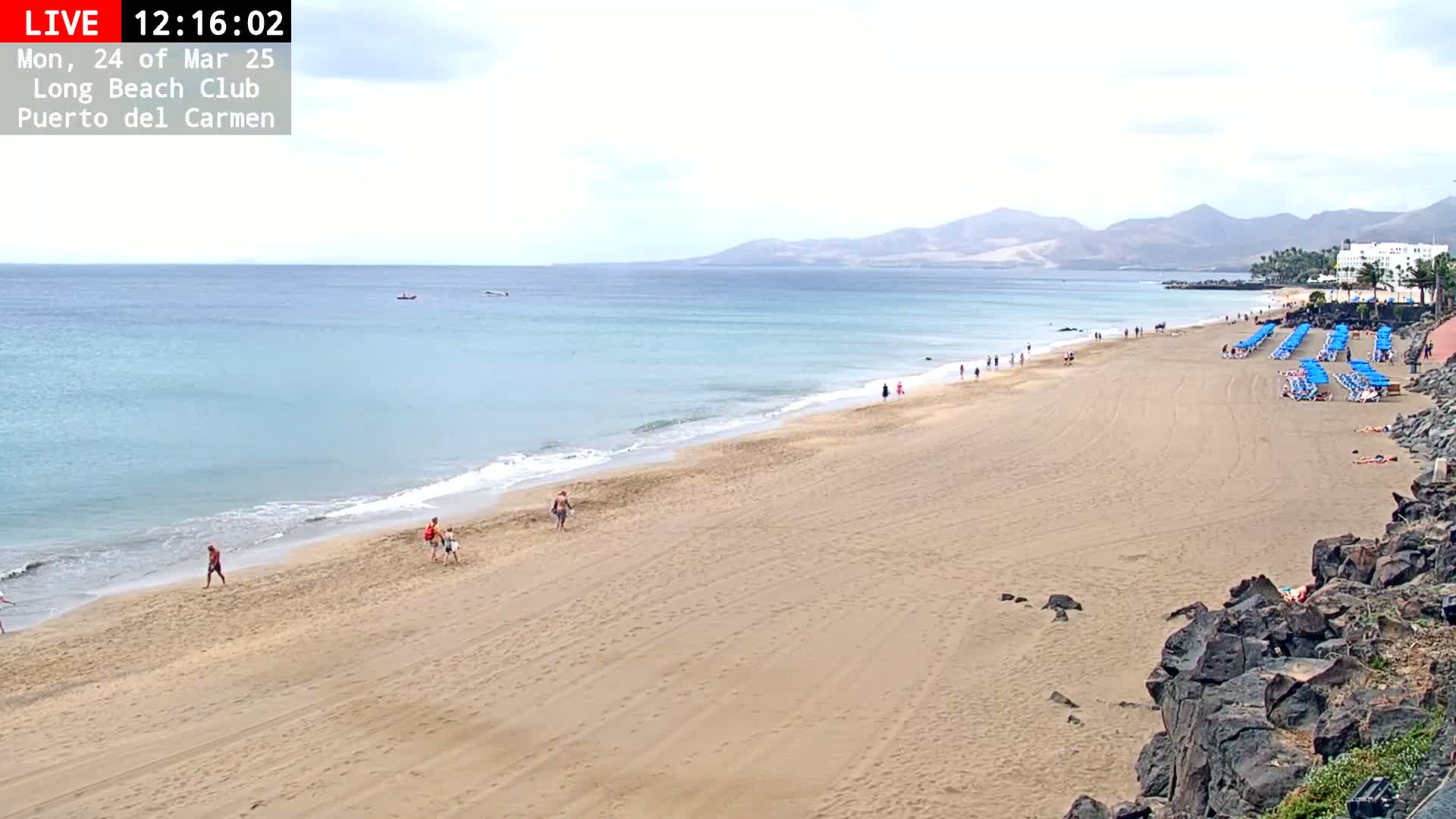 A sandy beach with a few people and rows of blue beach umbrellas stretches along a calm, light-blue ocean under a mostly cloudy sky, with mountains visible in the distance.