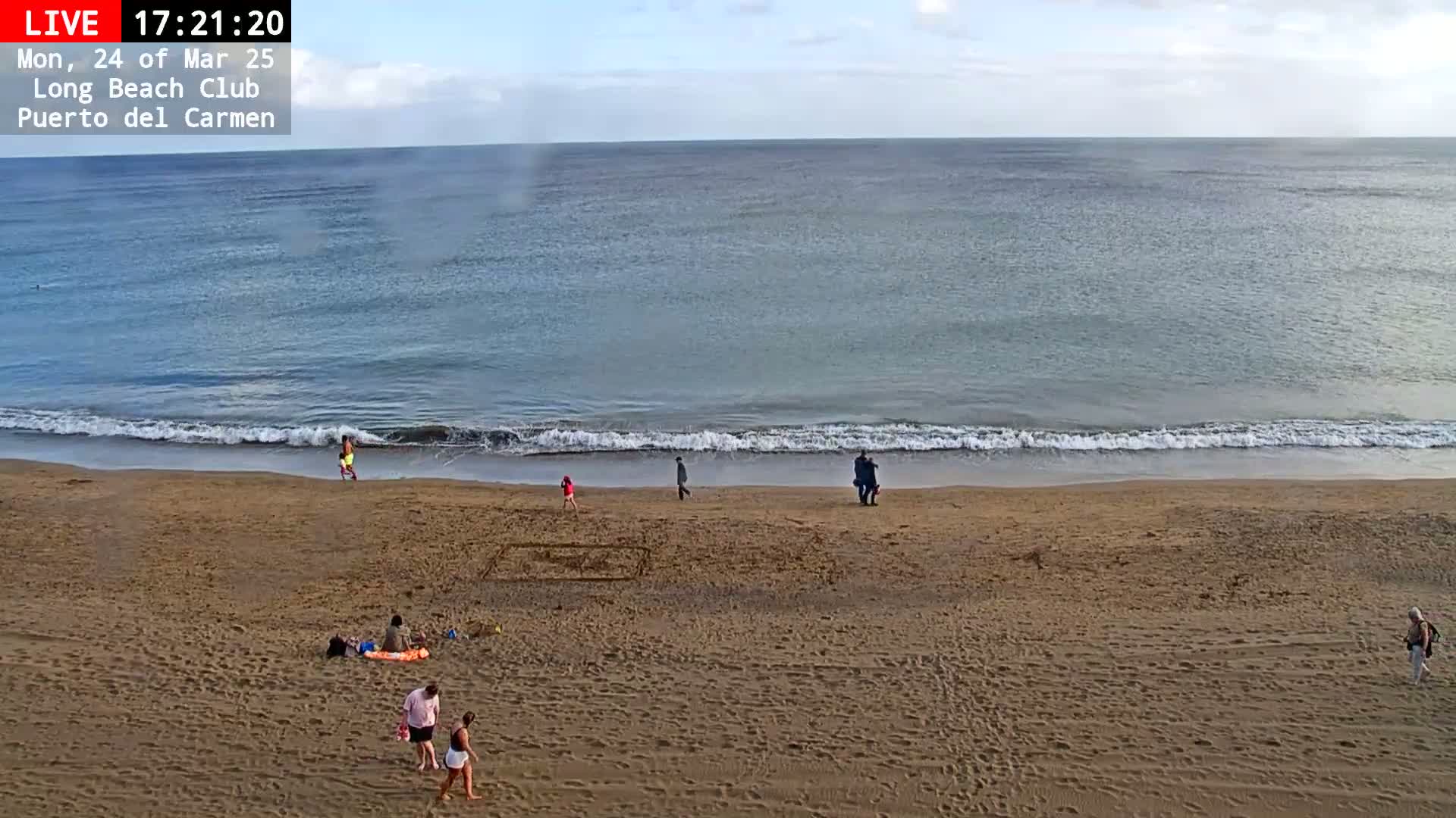 A sandy beach with a few people on it and gentle waves lapping the shore under a mostly clear sky.