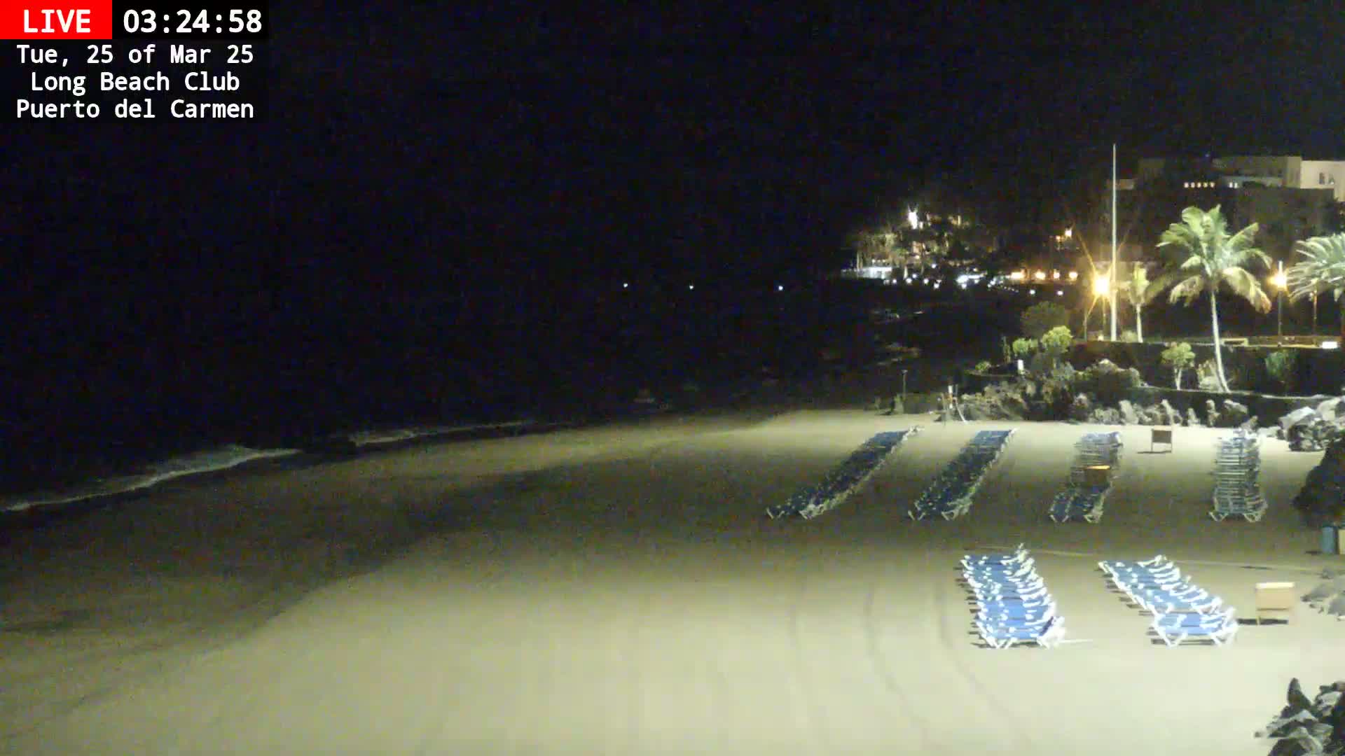 A nighttime view of a mostly empty sandy beach with rows of lounge chairs and a dark ocean with small waves under a clear, dark sky.
