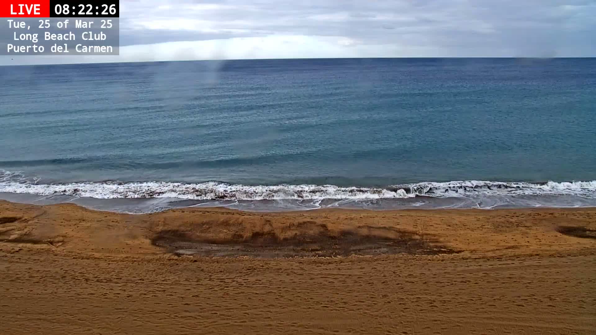 A sandy beach meets a calm, teal ocean under a cloudy sky.