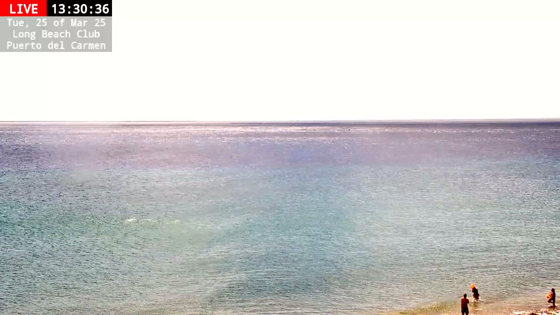 A calm, light-blue ocean stretches to a bright horizon under a clear sky, with a few people wading in the shallows near the shore.