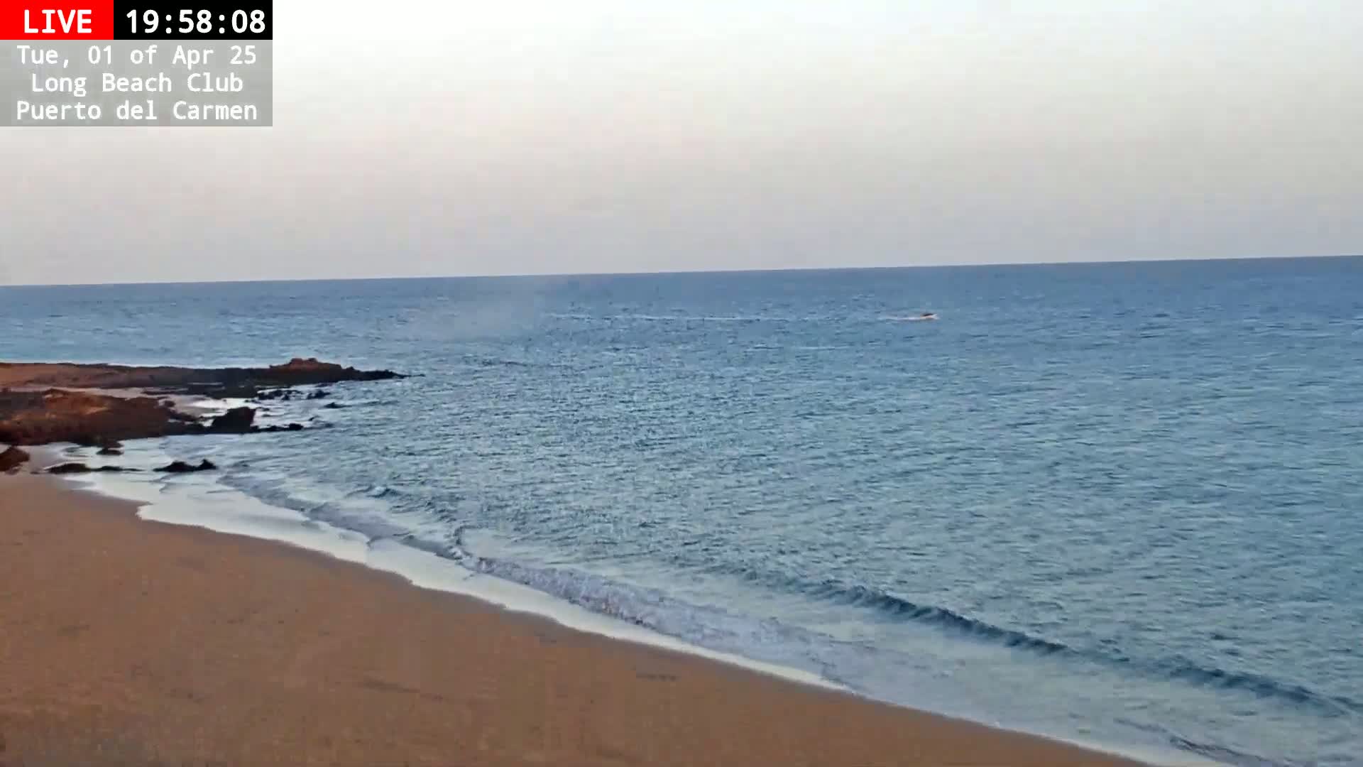 A sandy beach slopes gently down to a calm, blue ocean with small waves lapping the shore under a hazy sky.