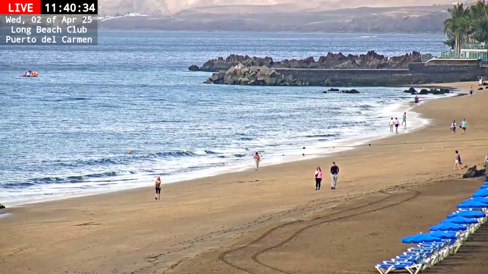A sandy beach with a few people walking along the shore, small waves lapping at the sand, and calm ocean waters under a hazy sky.