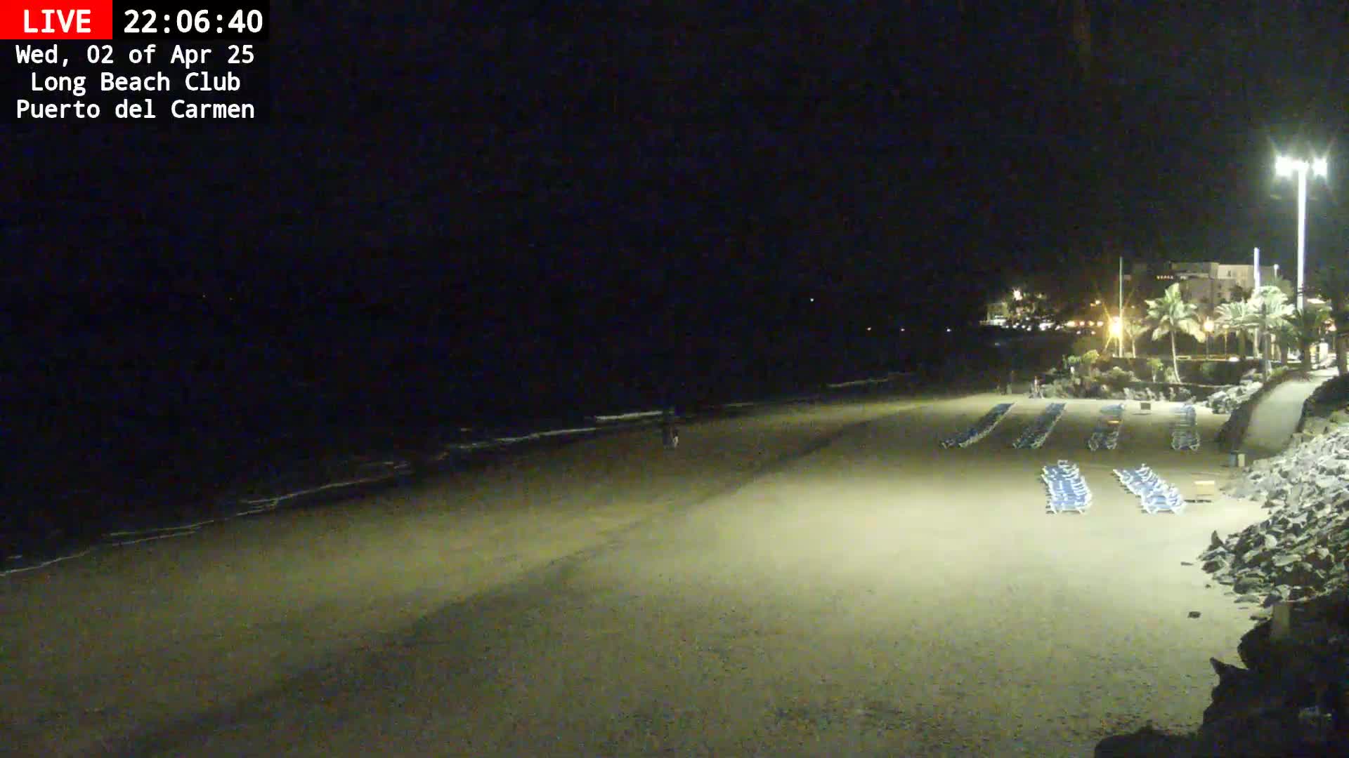 A dark sandy beach at night, with rows of empty lounge chairs and a calm ocean visible in the background under a clear sky.