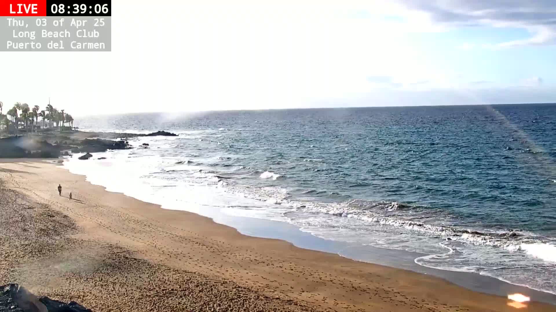 A sandy beach with two people walking along the shore, bordered by dark rocks and a calm ocean under a partly cloudy sky.