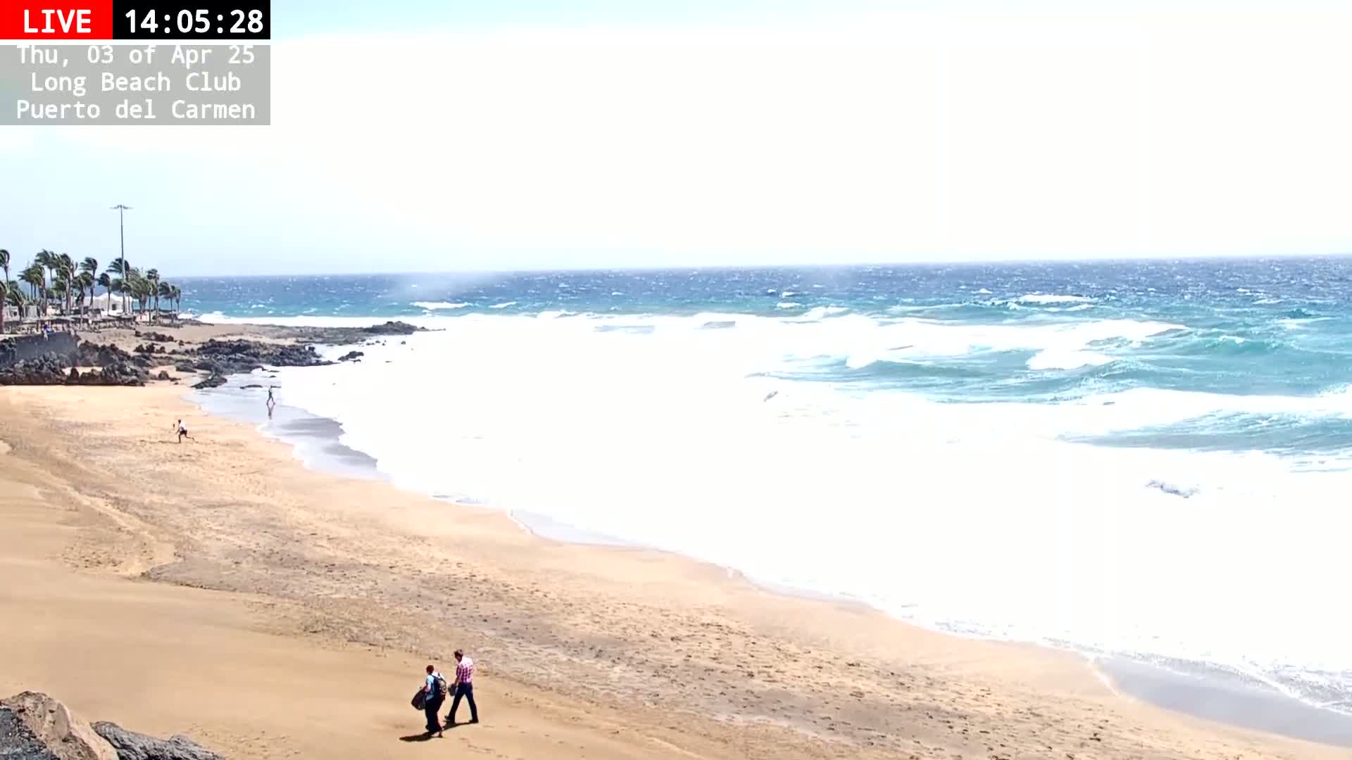 A sandy beach with a few people walking on it and moderately wavy ocean under a bright, sunny sky.