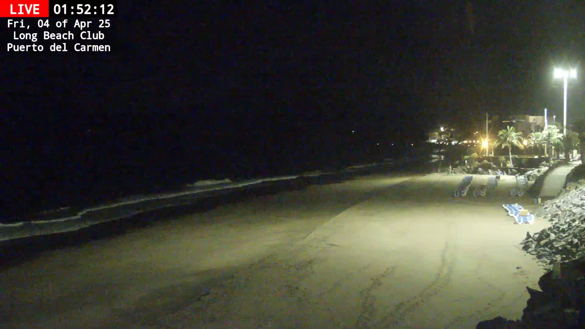 A mostly empty sandy beach at night is gently lapped by waves under a dark sky.