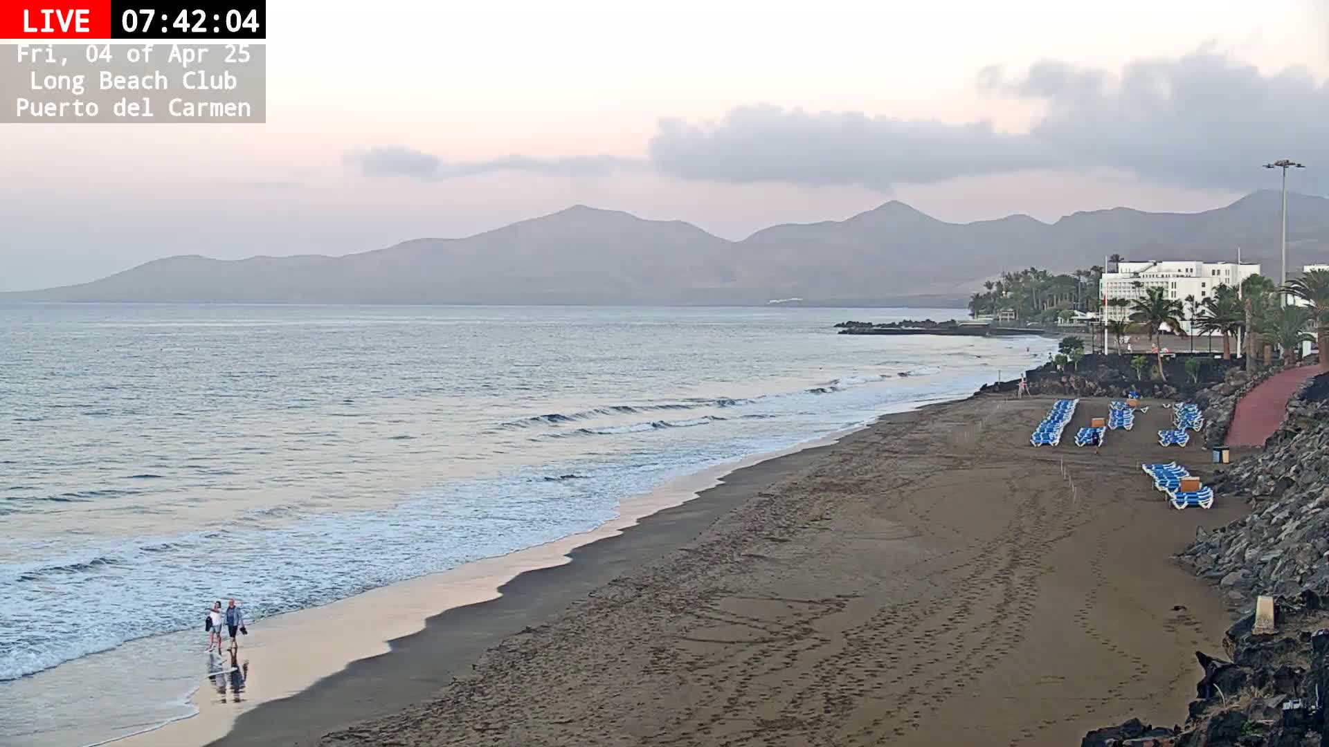 Two people walk along a sandy beach with calm waves under a cloudy sky, mountains visible in the distance.