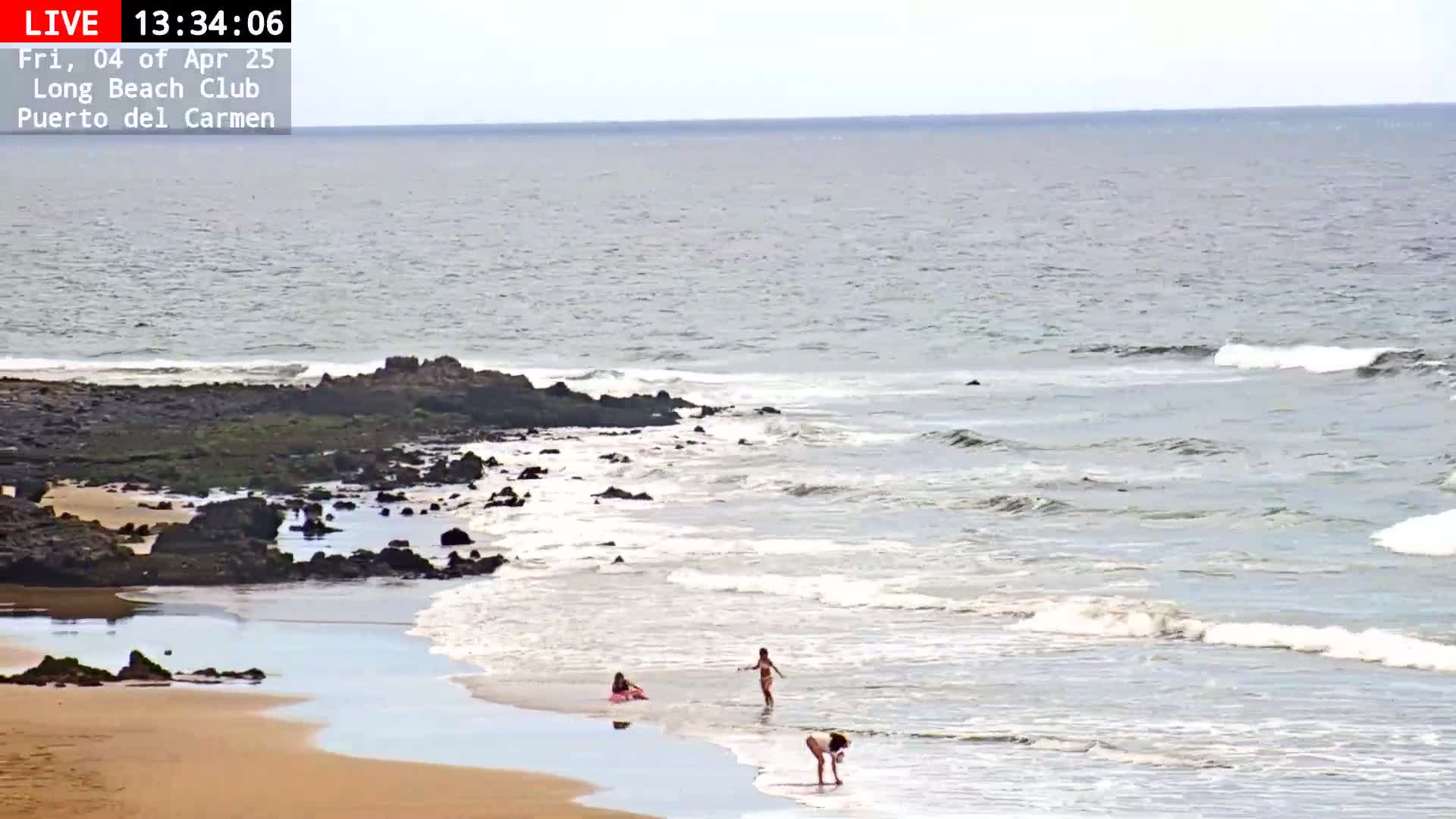 A sandy beach with dark rocks and calm, light gray waves laps the shore where several people are wading in the shallows under a mostly cloudy sky.