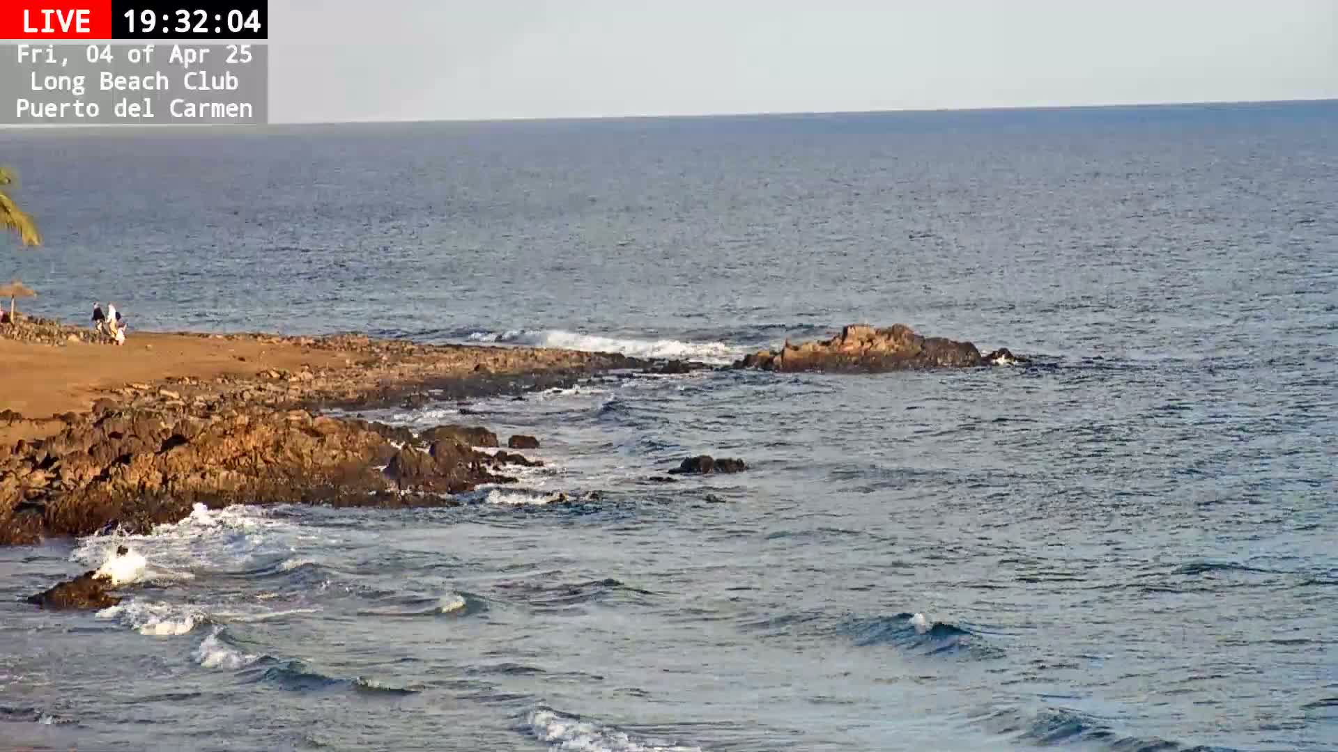 A rocky shoreline with a few people on a sandy beach meets a calm, blue ocean under a clear sky.