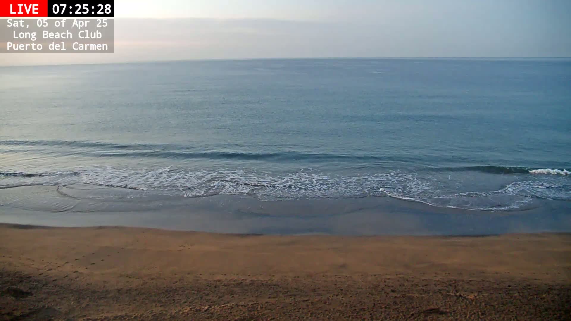 A sandy beach slopes down to a calm, blue ocean with small waves under a mostly clear sky.