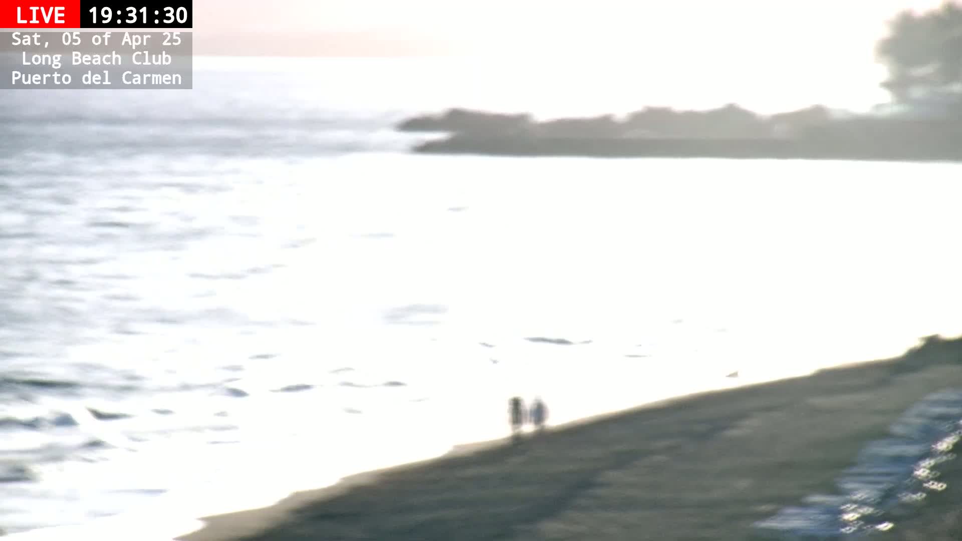 Two blurry figures walk along a sandy beach next to a calm, bright ocean under a mostly clear sky.