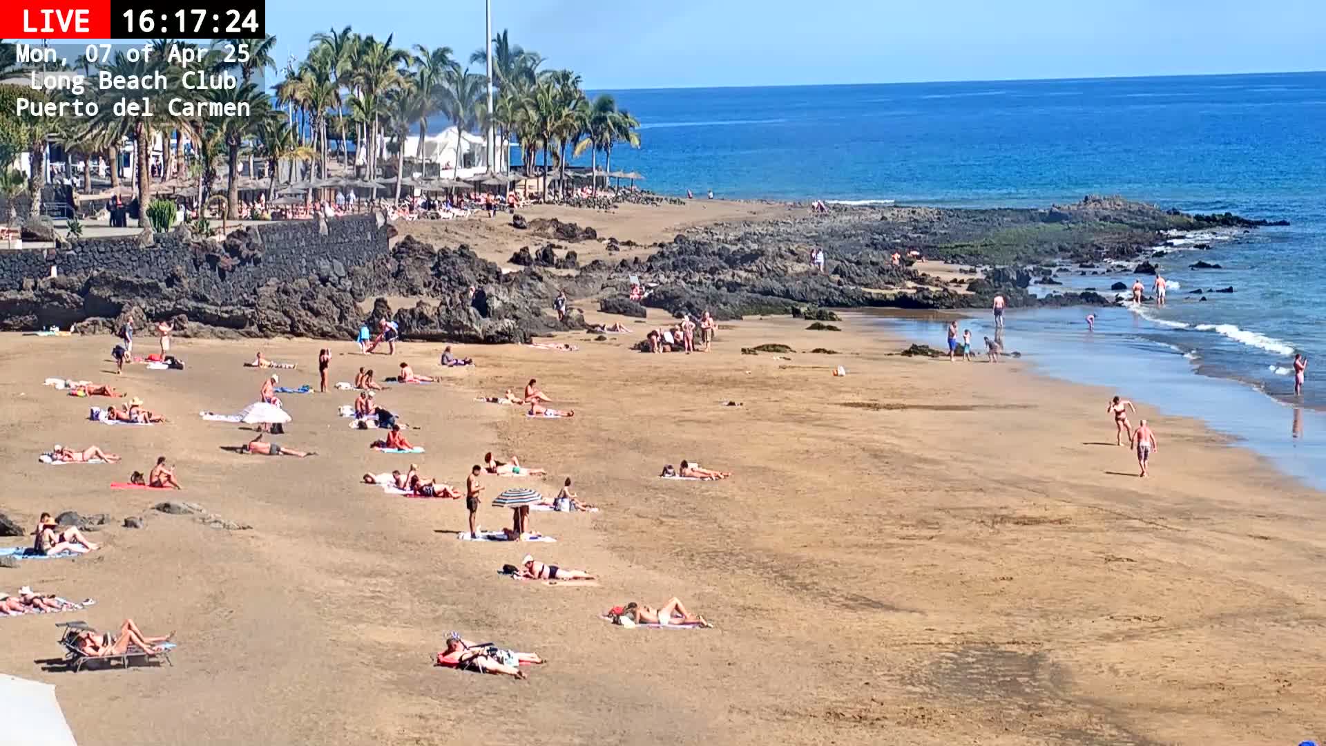 A sandy beach with many people sunbathing and swimming in a calm ocean under a sunny sky.