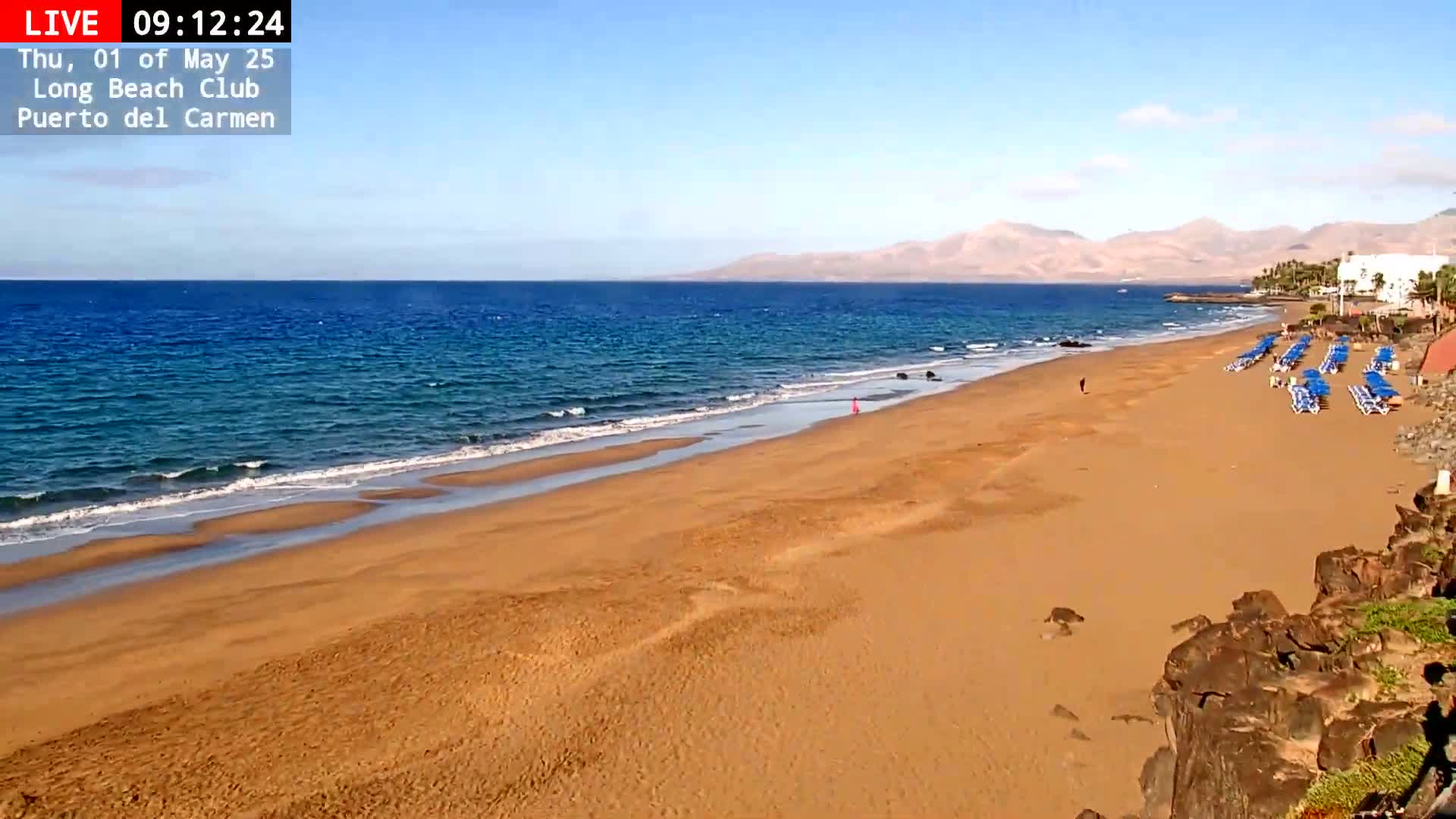 A long sandy beach with calm blue water stretches out towards distant mountains under a clear blue sky.