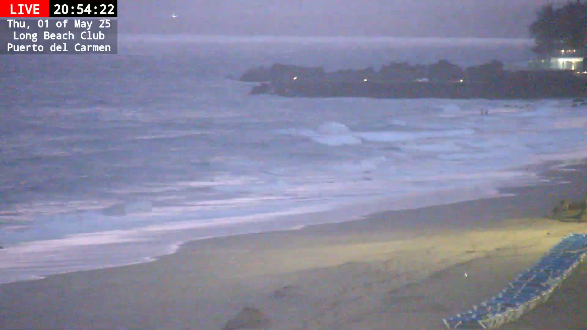 A dark, overcast evening shows a beach with moderate waves rolling onto the shore.