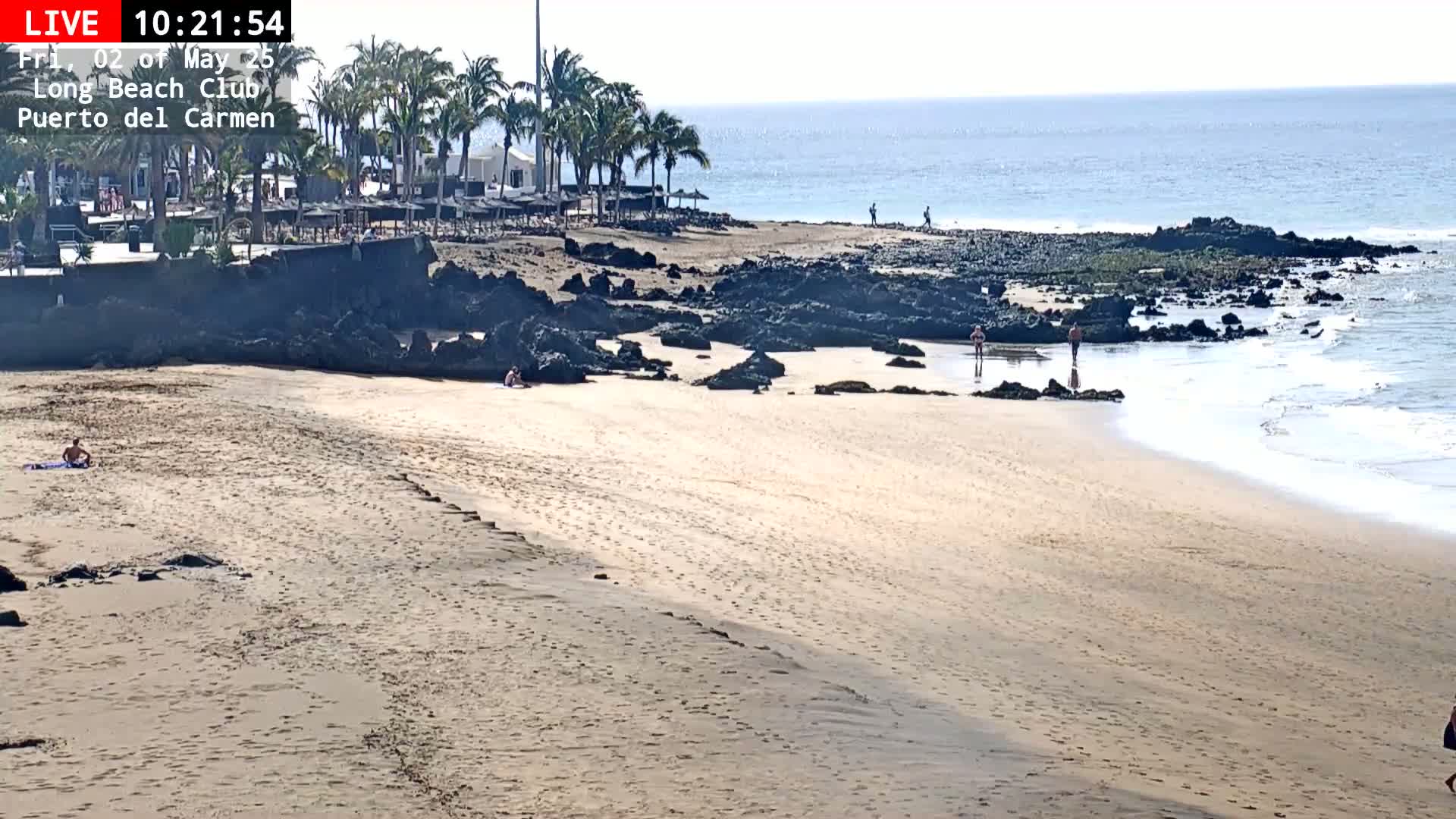 A sandy beach with a few people on it, some palm trees, dark rocks, and a calm ocean under a sunny sky.