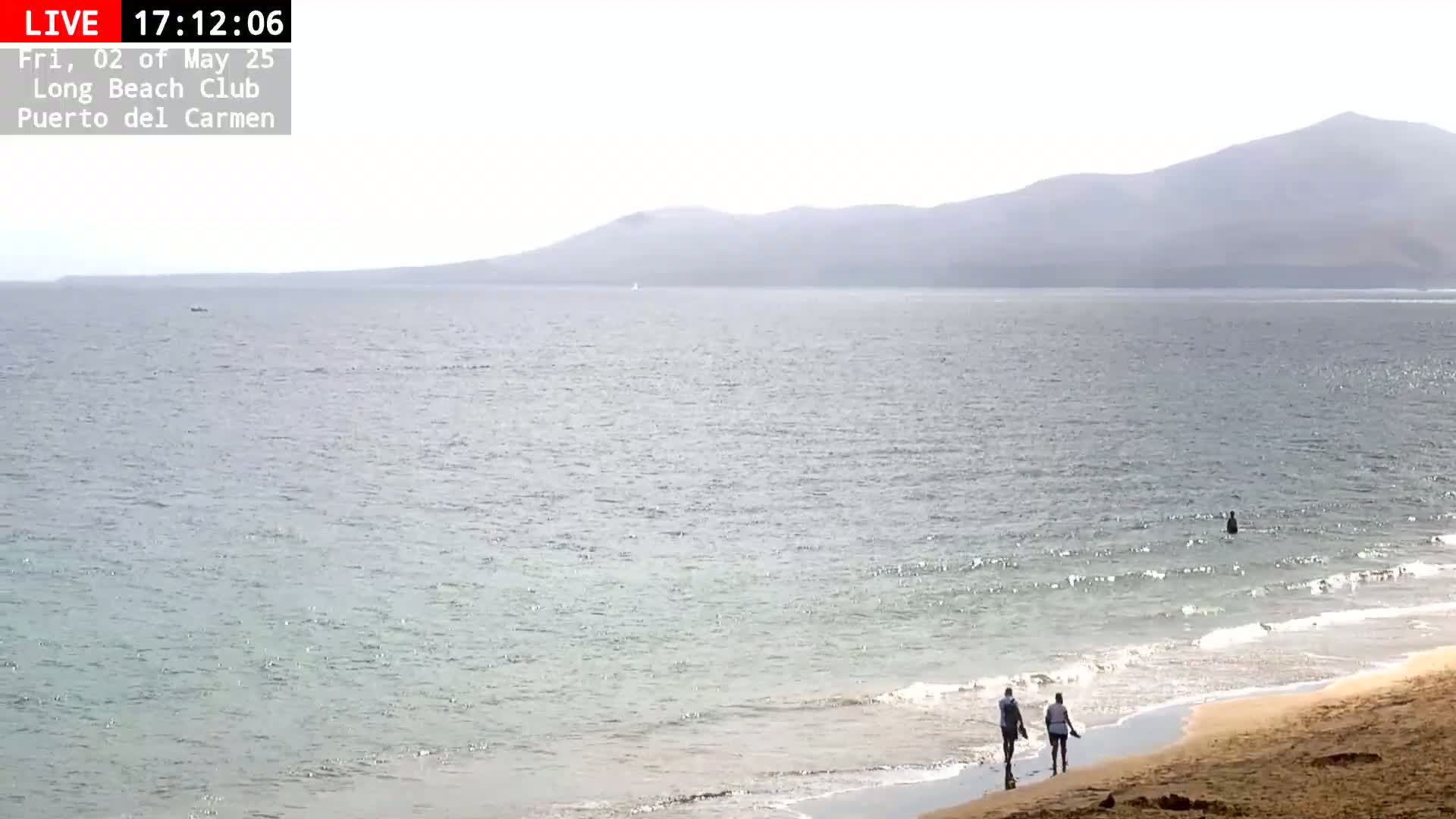A calm, sunny ocean beach scene shows two people walking on the sand near the water's edge, with a hazy mountain range in the distance.