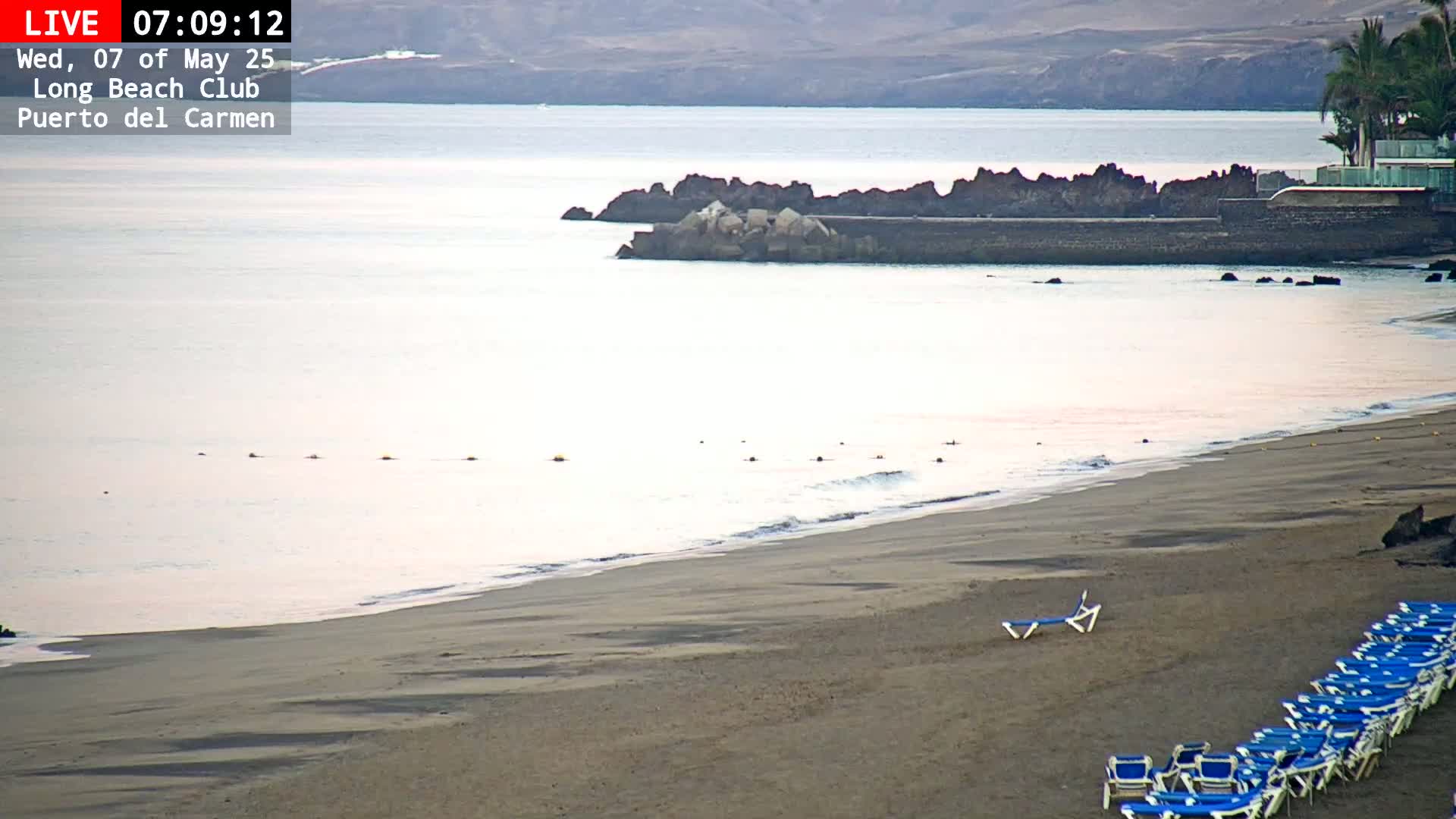 A sandy beach with calm, light-colored water and several rows of empty blue lounge chairs is visible under a hazy sky.