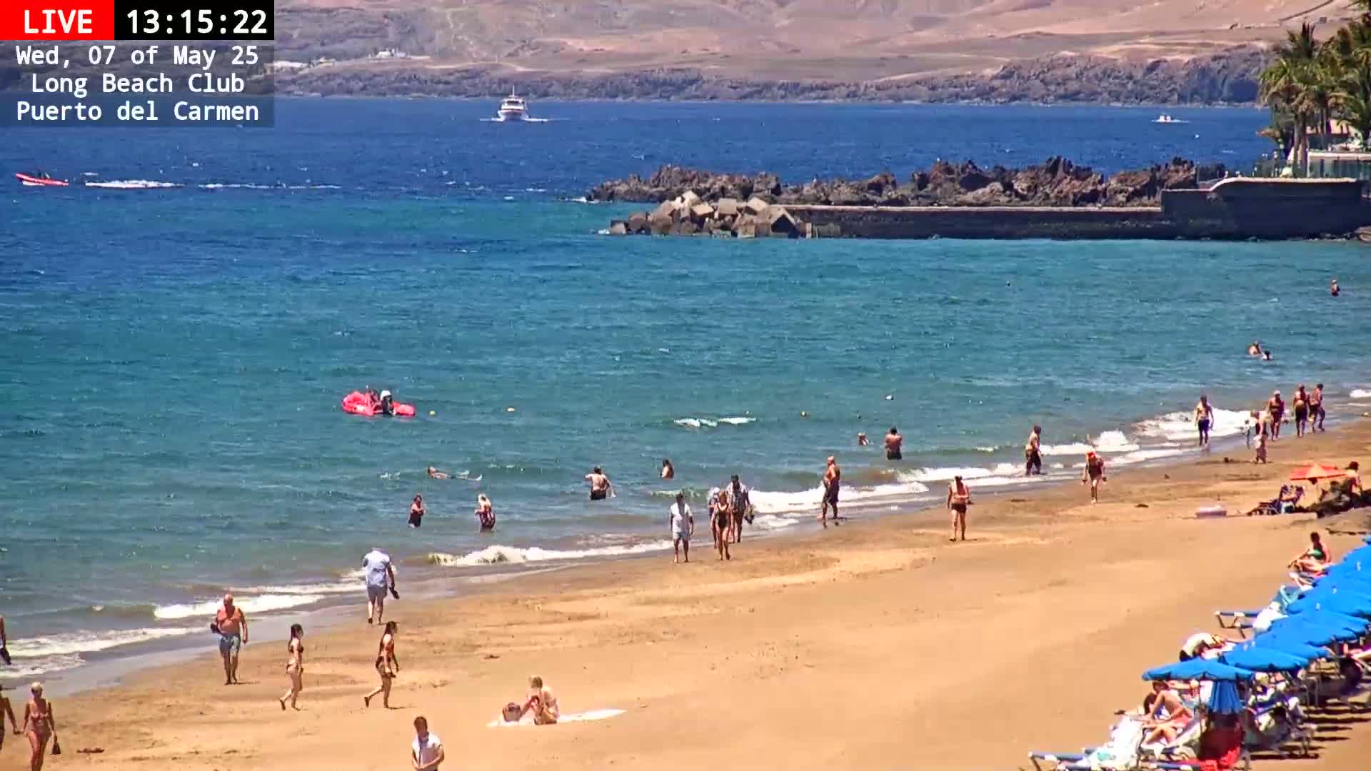A sandy beach with many people swimming, sunbathing, and walking along the shore, under a sunny sky; in the background, calm, clear blue water extends to a distant shoreline.