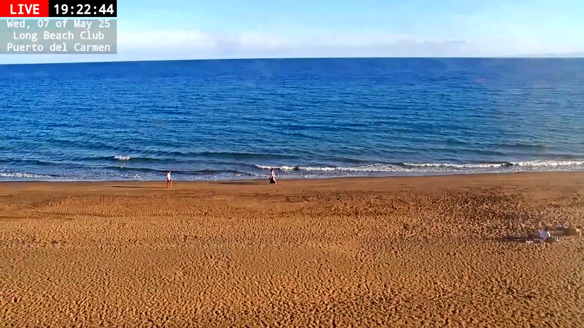 A wide sandy beach with a few people walking and sitting on it meets a calm, blue ocean under a clear sky.