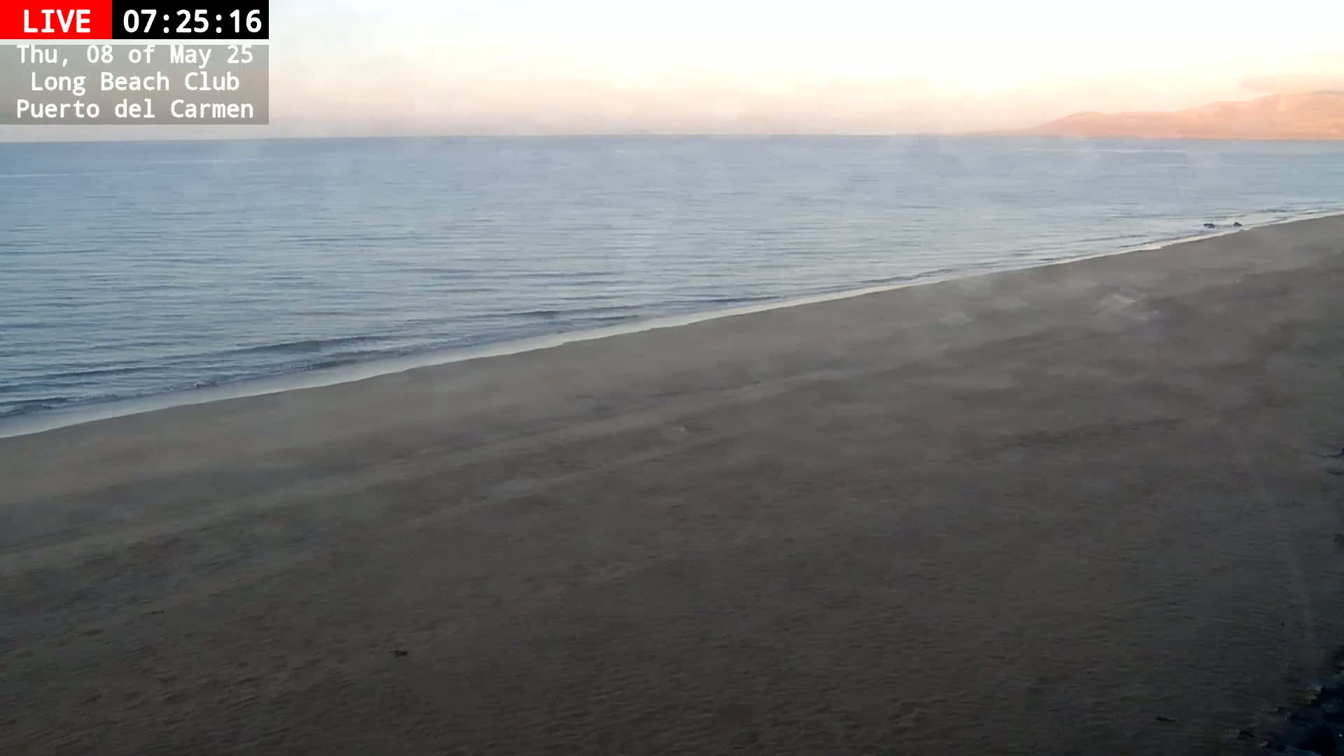 A long, sandy beach meets a calm, hazy ocean under a pale, sunrise sky.