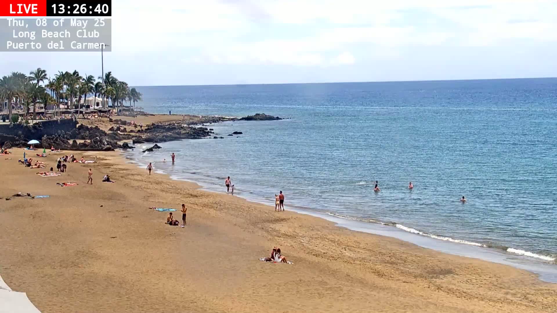 A sandy beach with numerous people sunbathing and swimming in calm, clear blue ocean water under a partly sunny sky.