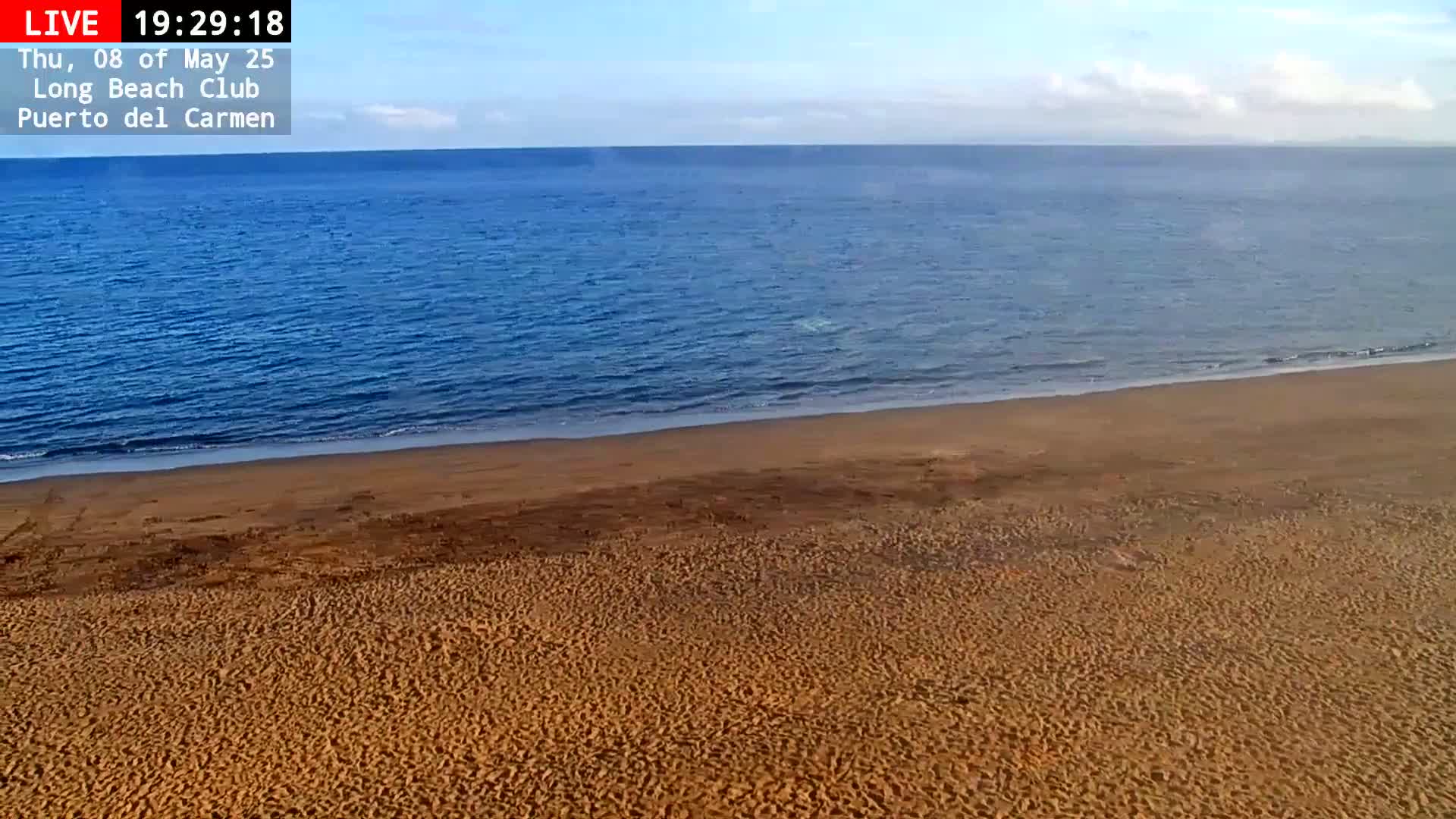 A mostly clear sky overlooks a calm blue ocean meeting a wide, sandy beach.