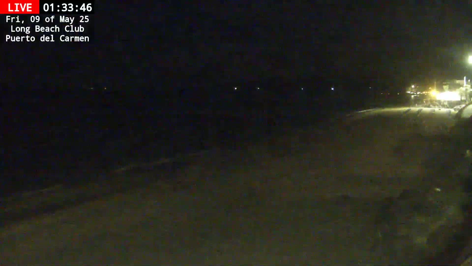 A dark, nighttime view of a beach with dimly lit structures in the distance.