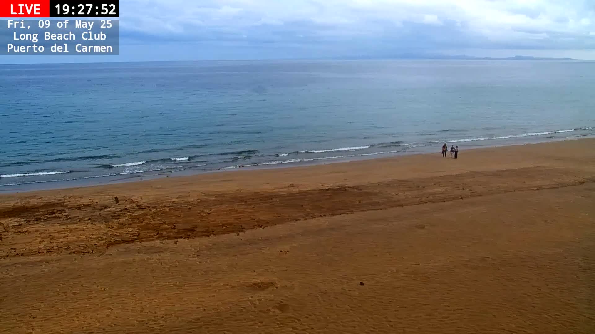 A mostly sunny day shows a wide sandy beach with a few people walking near the water's edge, and a calm ocean with small waves in the background.