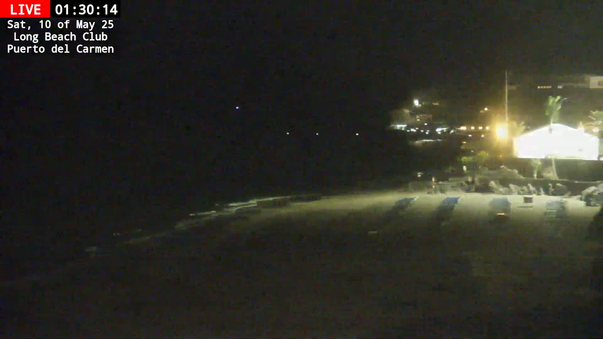A dark, nighttime beach scene shows an empty sandy shore with several beach chairs and a building lit up in the background under a clear, starlit sky.