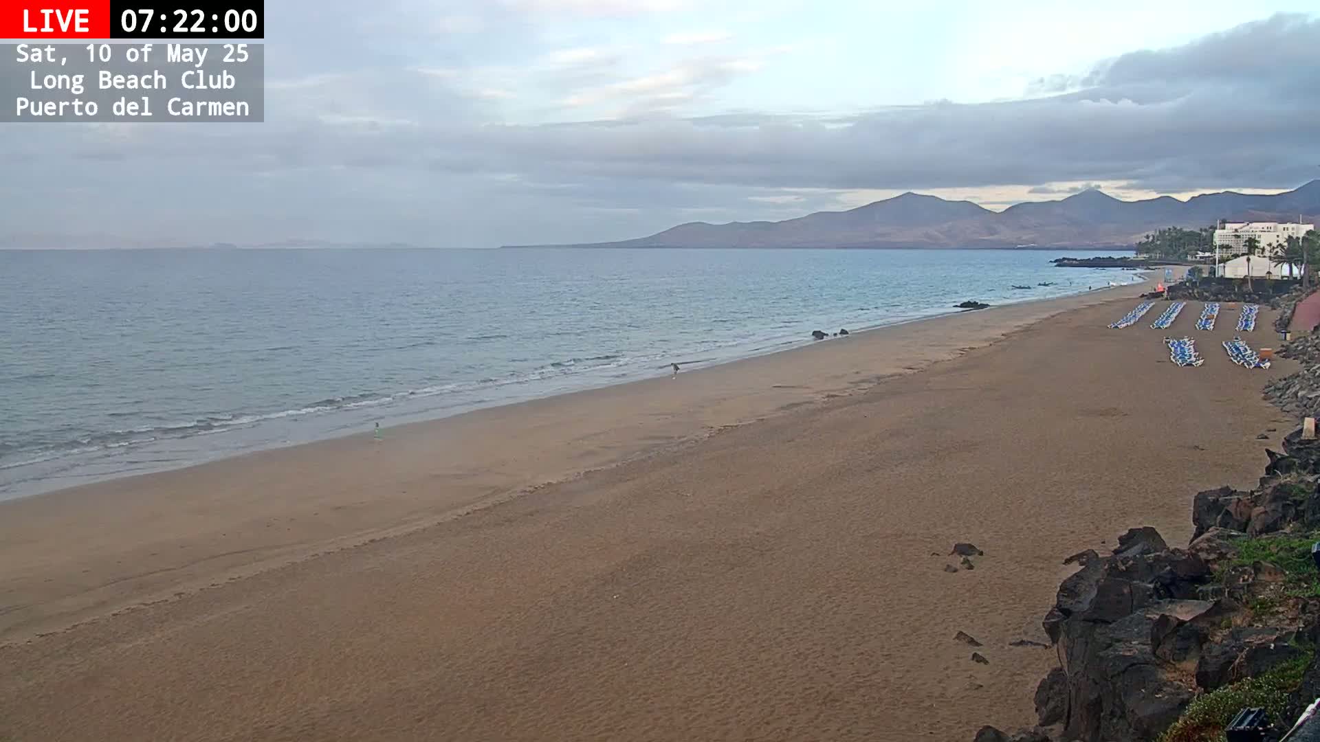 A long, sandy beach stretches towards a calm ocean under a mostly cloudy sky, with distant mountains visible in the background.
