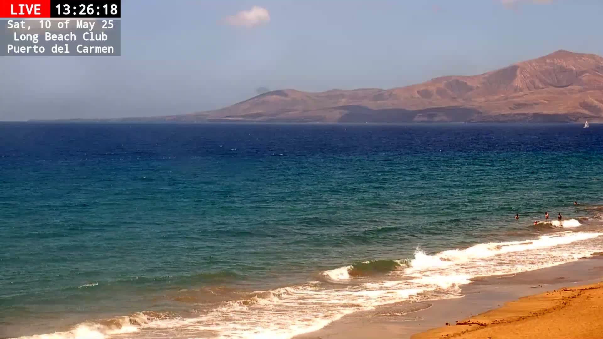 A sandy beach with small waves laps the shore, under a clear sky, with a distant mountain range visible across a calm ocean.
