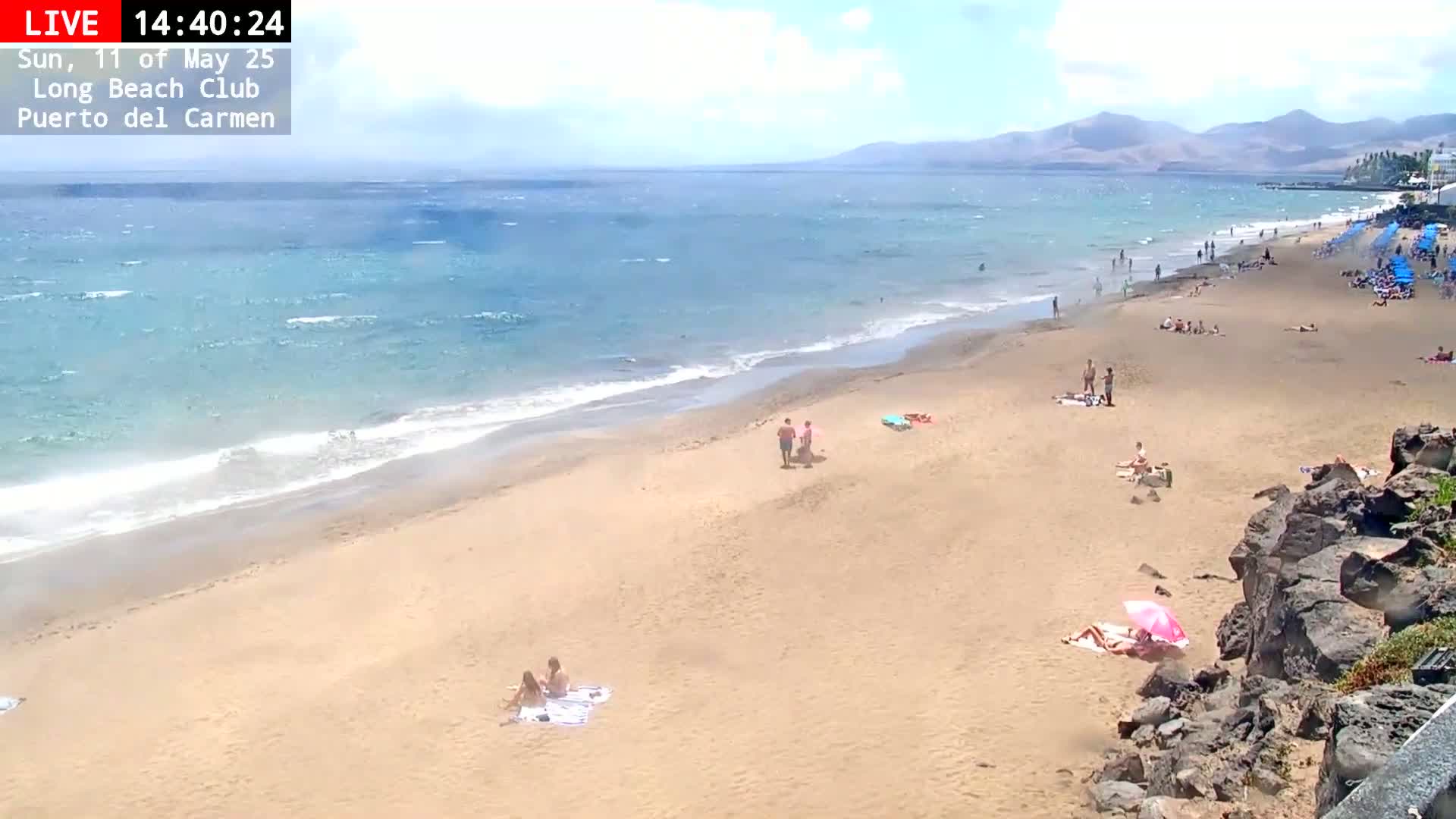 A sandy beach with a moderate number of people sunbathing and swimming, under partly cloudy skies, with a gently waving ocean in the background.