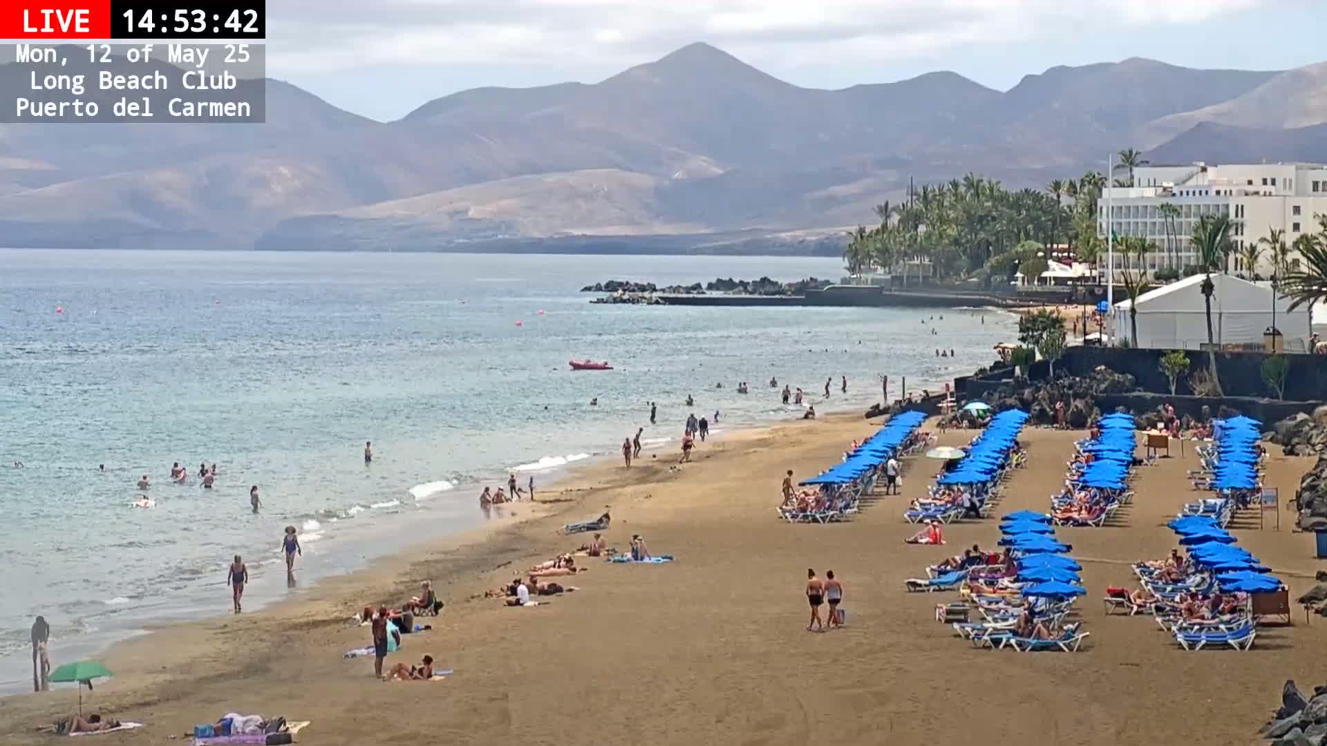 A sandy beach with many people swimming and sunbathing under blue umbrellas, backed by a calm ocean, mountains, and a hotel under a partly cloudy sky.