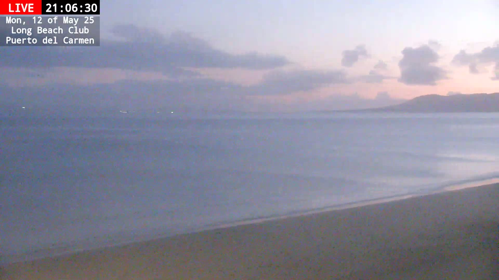 A sandy beach meets a calm, dusky ocean under a partly cloudy sky.