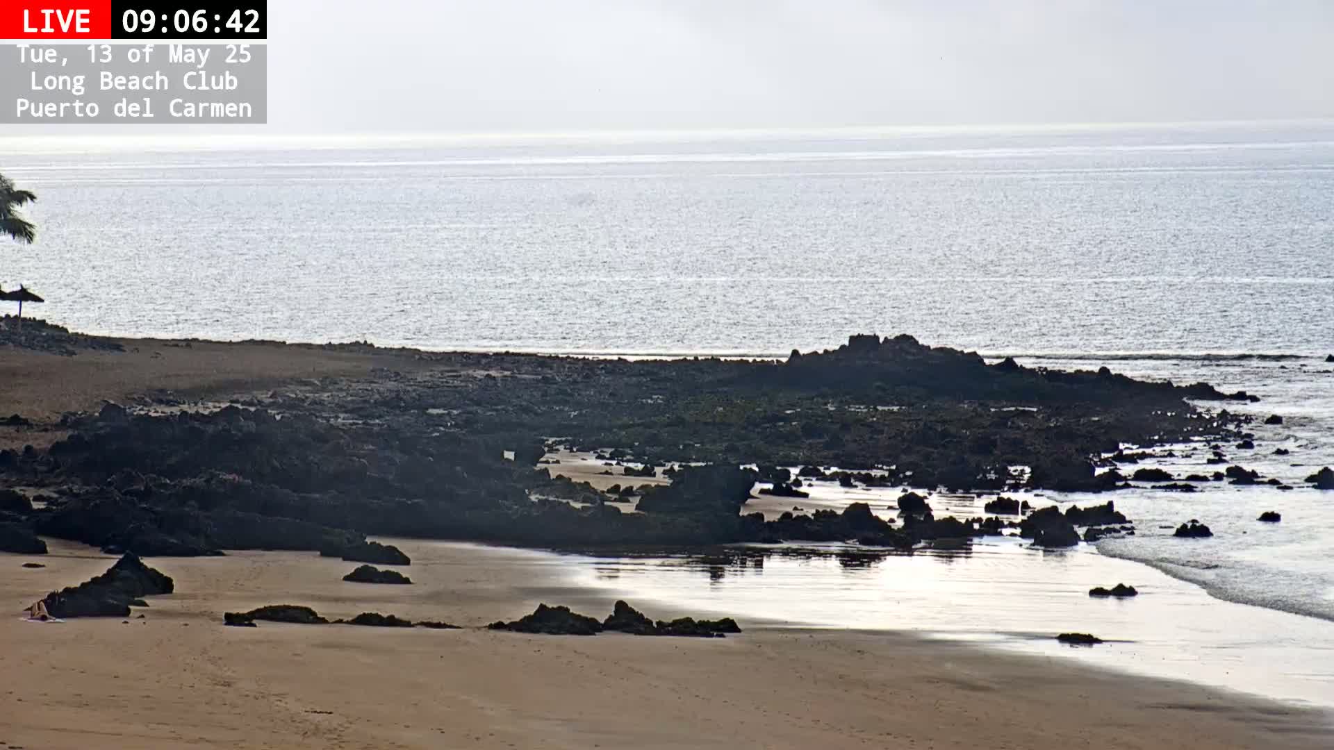 A sandy beach with dark rocks leading into a calm, light-grey ocean under a mostly clear sky.