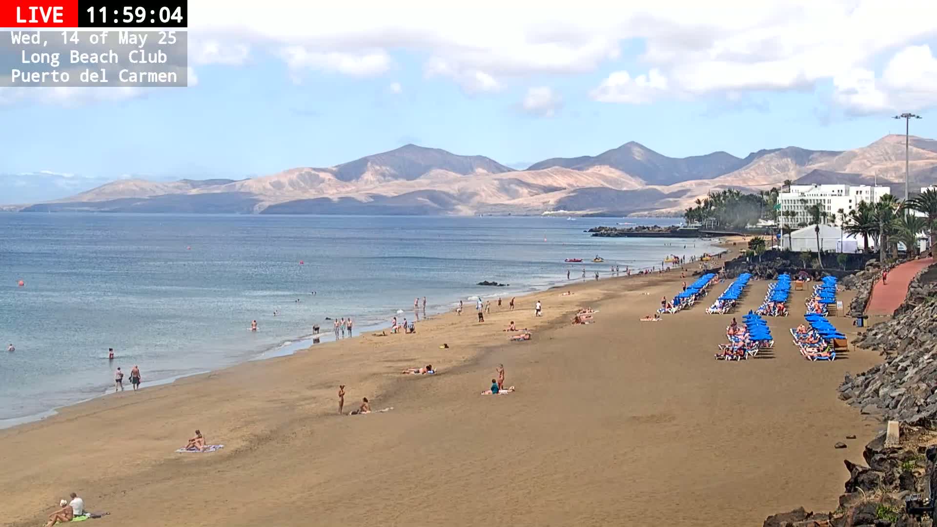 A sandy beach with people swimming and sunbathing, rows of blue umbrellas and lounge chairs, and a calm ocean with mountains in the background under a partly cloudy sky.