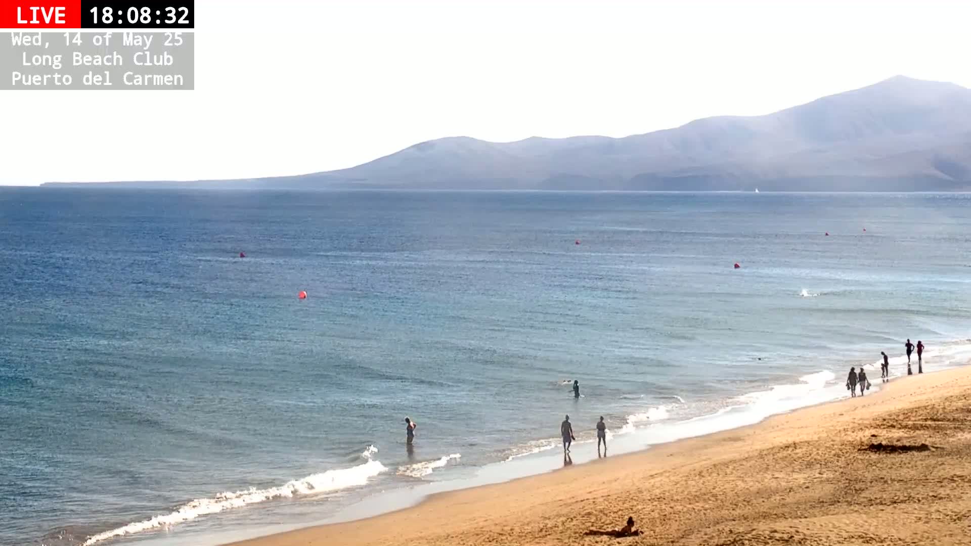 A sandy beach with several people wading in the calm, blue ocean under a bright, sunny sky, with distant mountains visible on the horizon.
