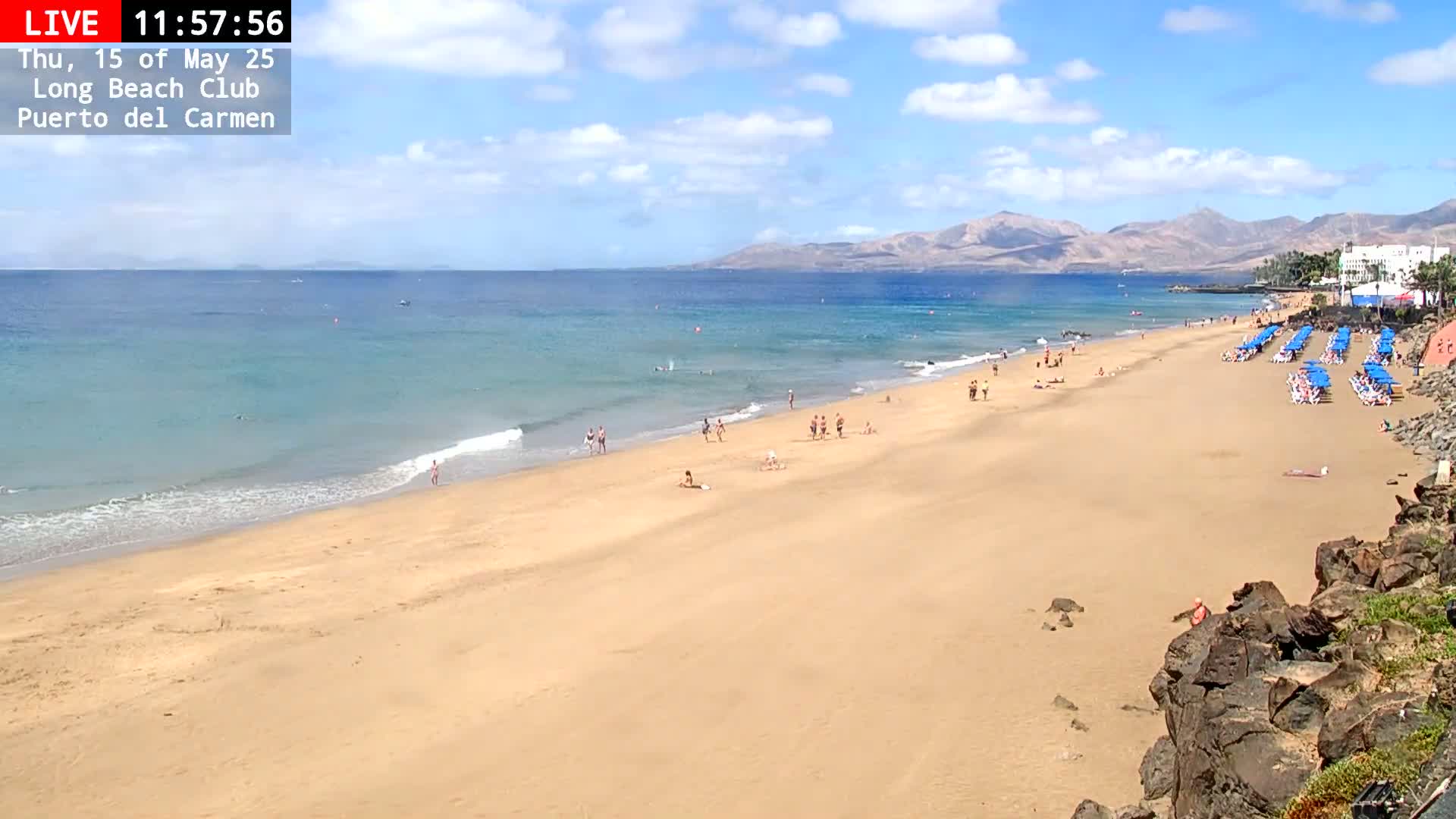 A mostly sunny day at a sandy beach with calm ocean waves, people sunbathing and swimming, and rows of blue beach umbrellas.