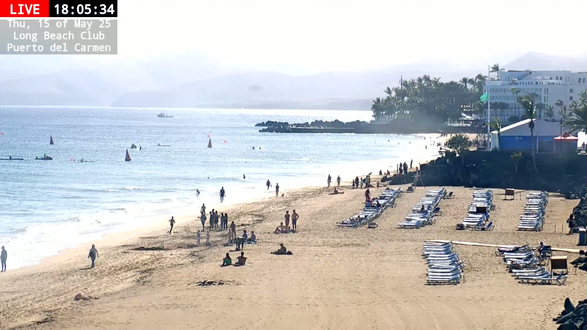 A sandy beach with people sunbathing and swimming in a calm ocean under a hazy sky, with rows of empty lounge chairs and a hotel in the background.