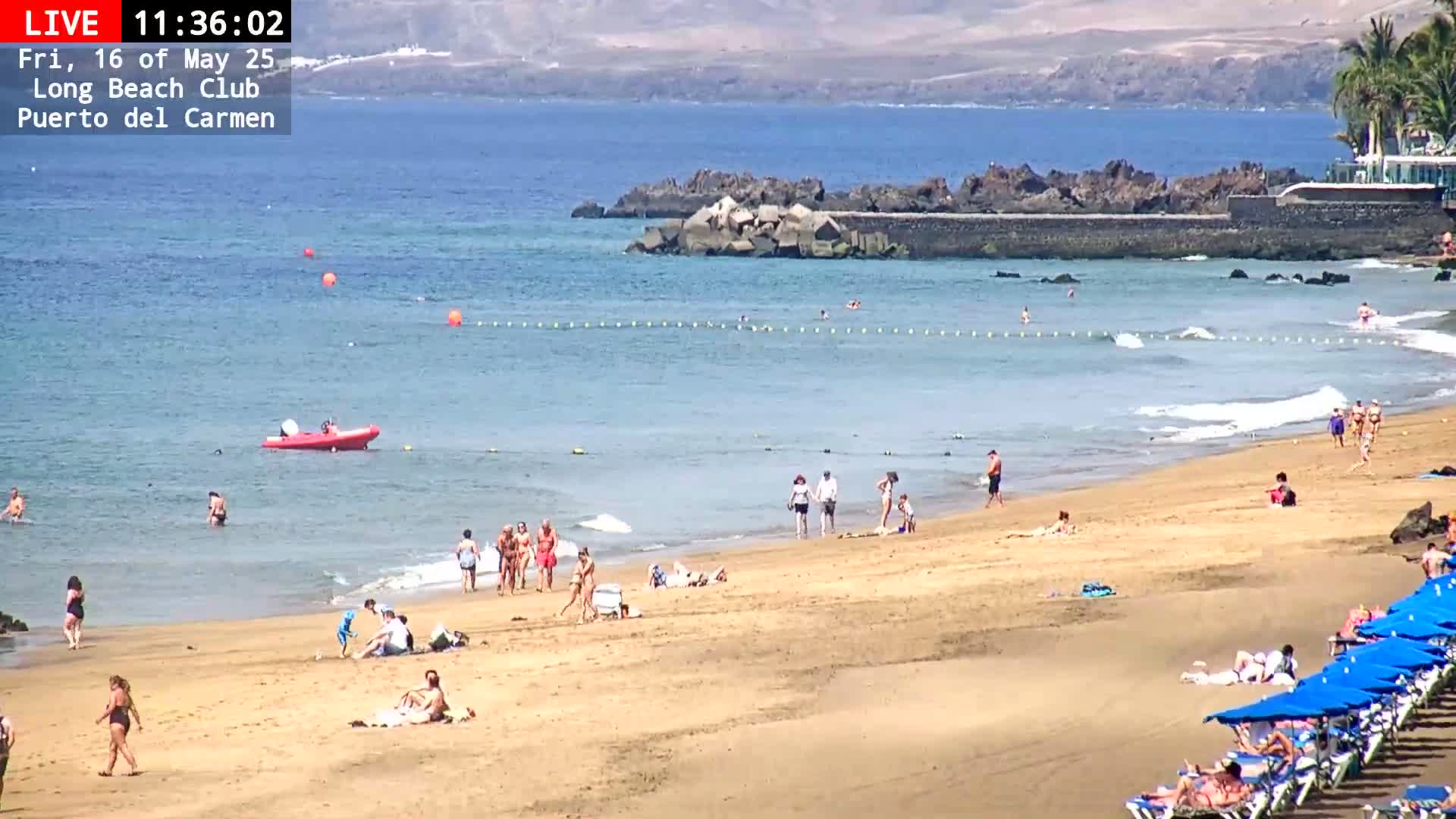 A sandy beach with numerous people swimming and sunbathing, under a sunny sky, with calm ocean waves gently lapping the shore.