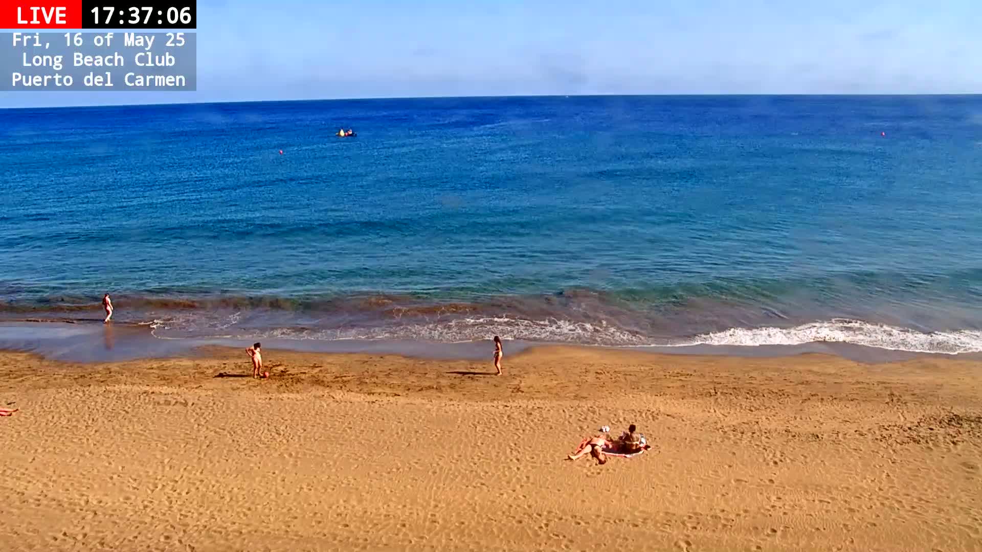 A sandy beach with a few people on it is next to a calm, blue ocean under a clear sky.