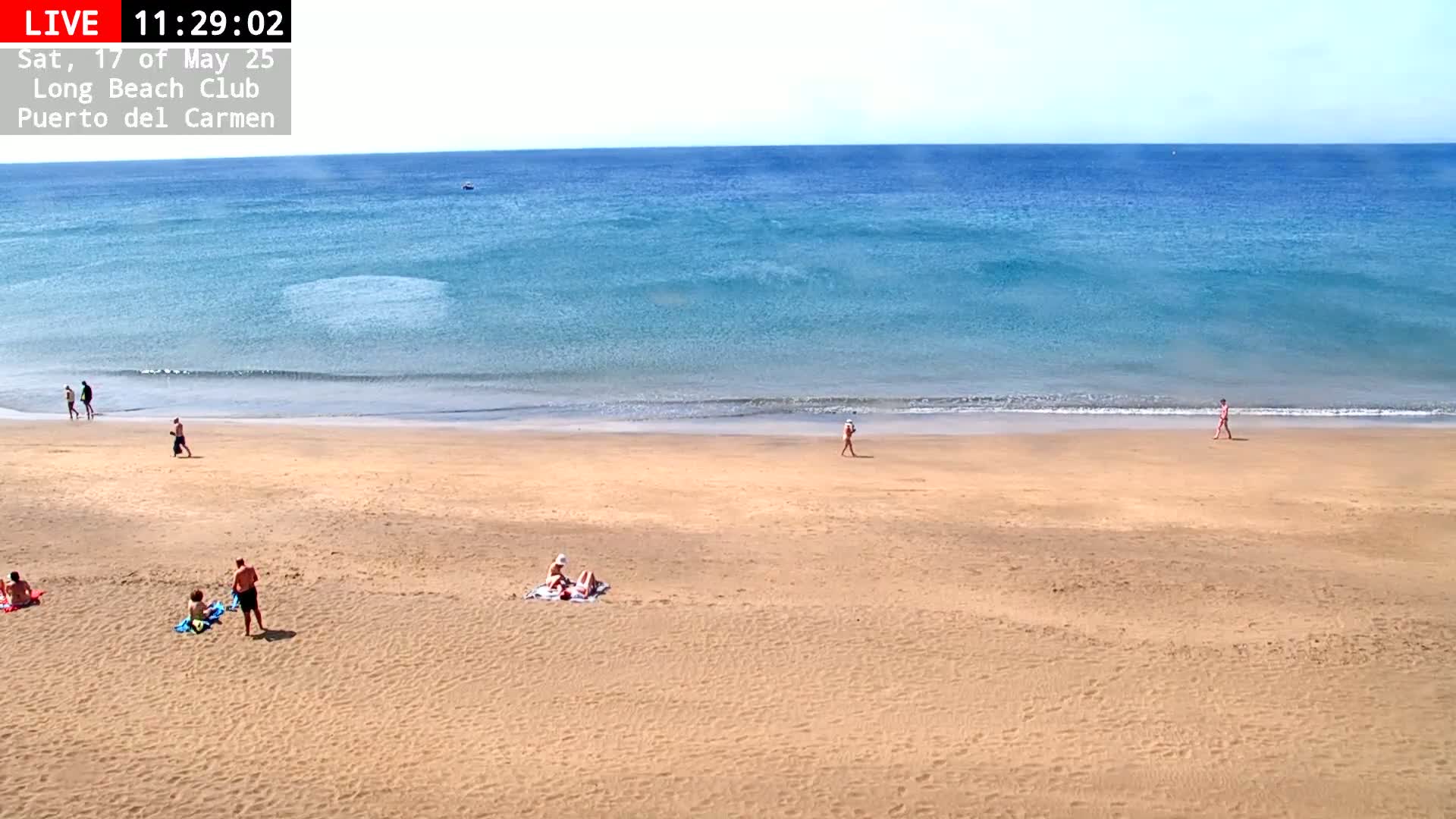 A sandy beach with a few people sunbathing and walking, overlooking a calm, blue ocean under a clear sky.