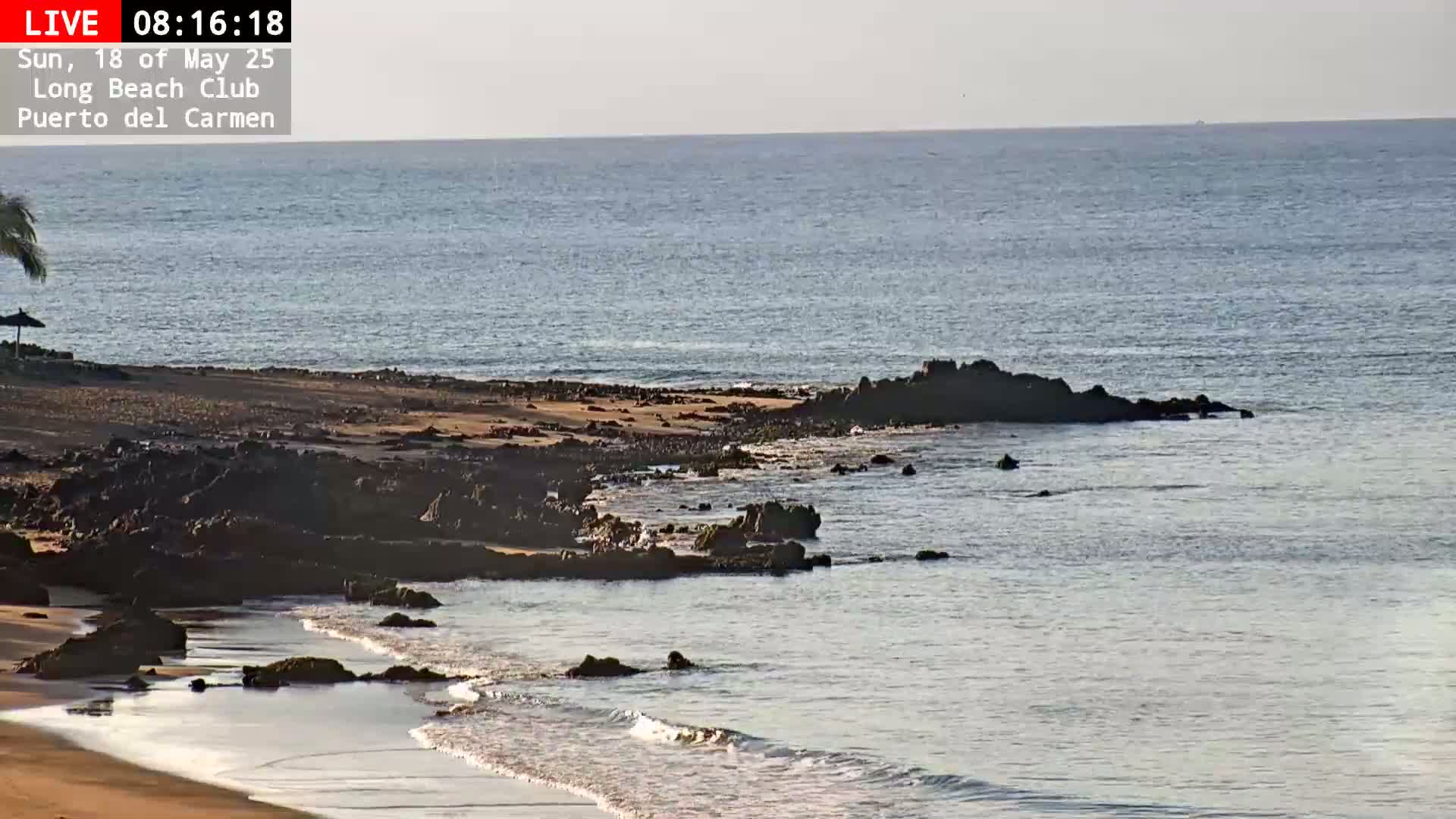 A sandy beach with dark rocks extends into a calm, light-blue ocean under a clear sky.