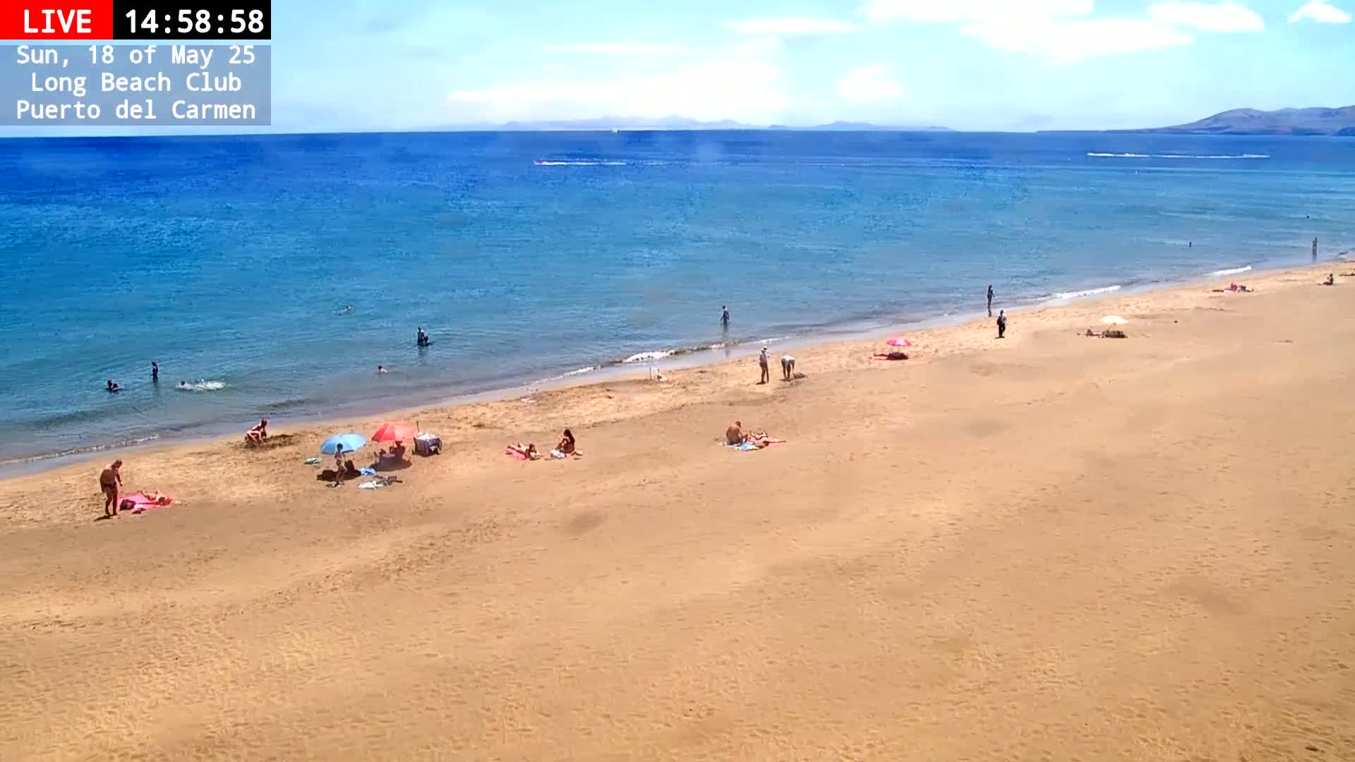 A sandy beach with scattered people sunbathing and swimming in a calm, blue ocean under a mostly sunny sky.