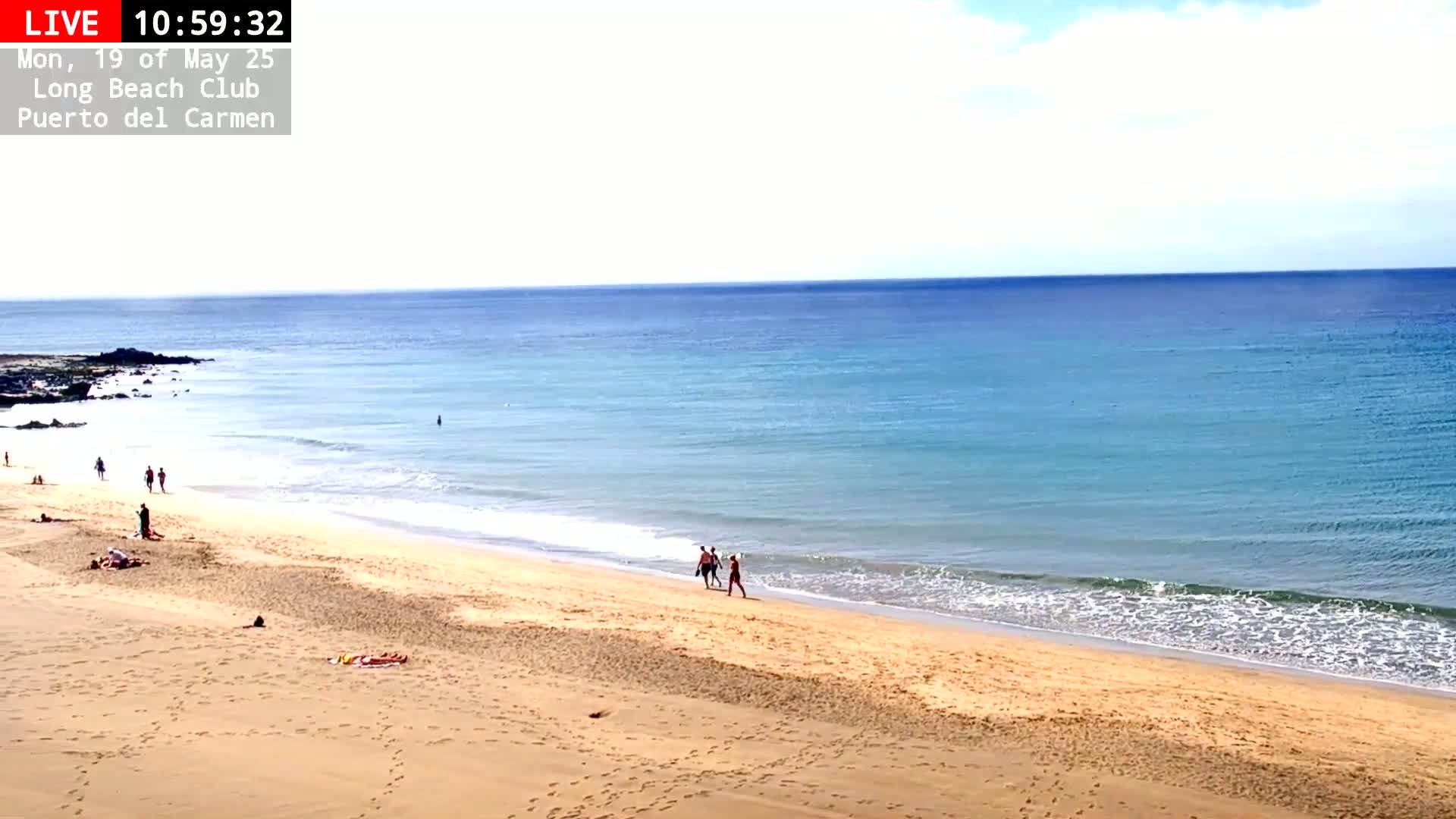 A sandy beach with a few people on it slopes down to a calm, light blue ocean under a mostly clear sky.