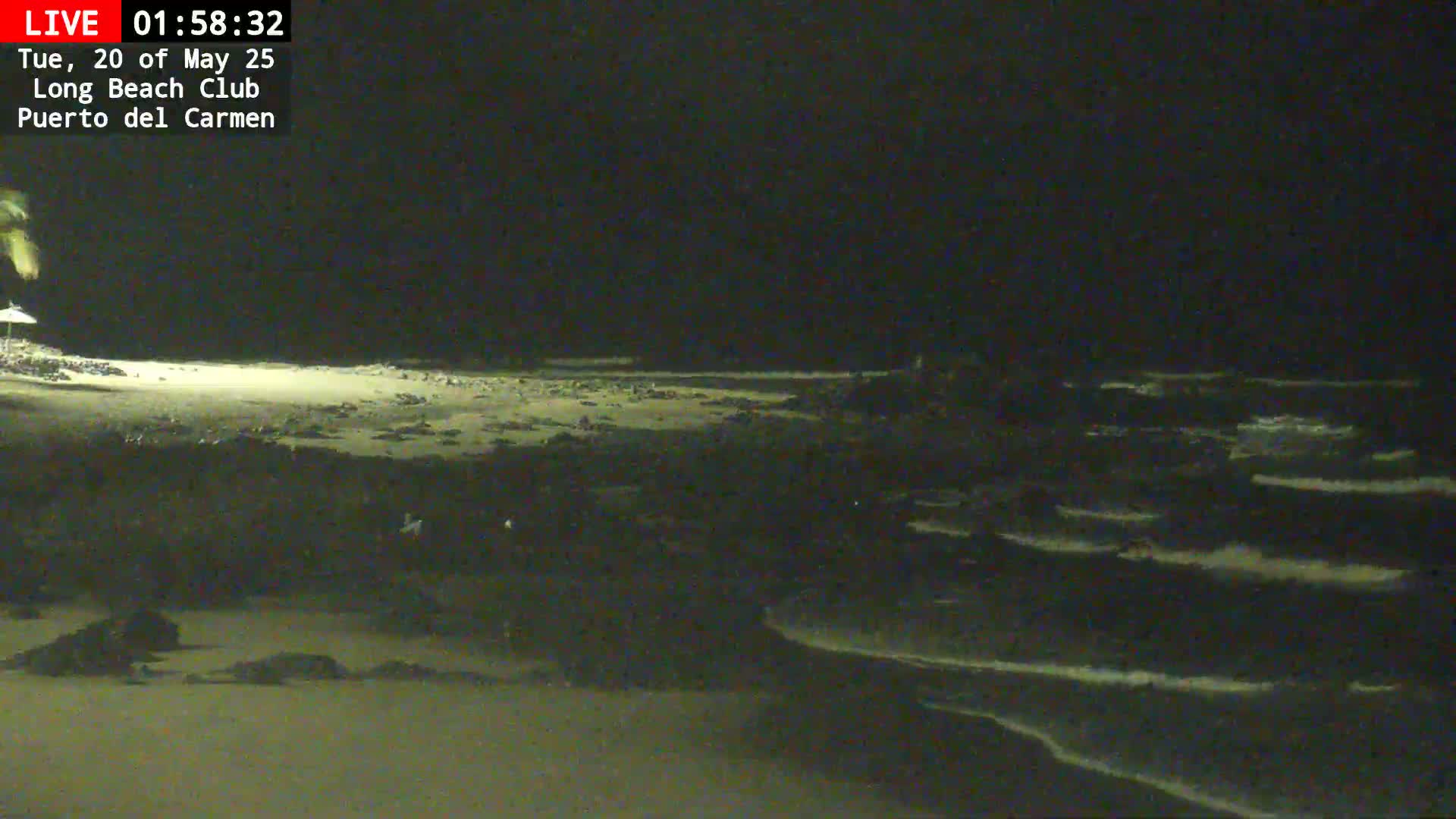 A dark, nighttime beach scene shows gently lapping waves on a sandy shore with some rocks visible near the waterline.