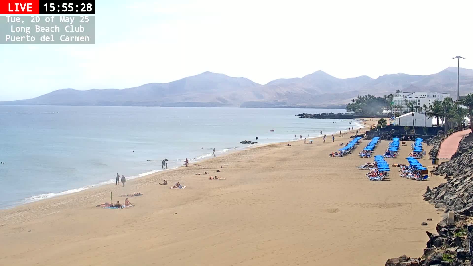 A sandy beach with scattered people sunbathing and swimming in calm ocean water, under a mostly clear sky, backed by distant mountains and a hotel.