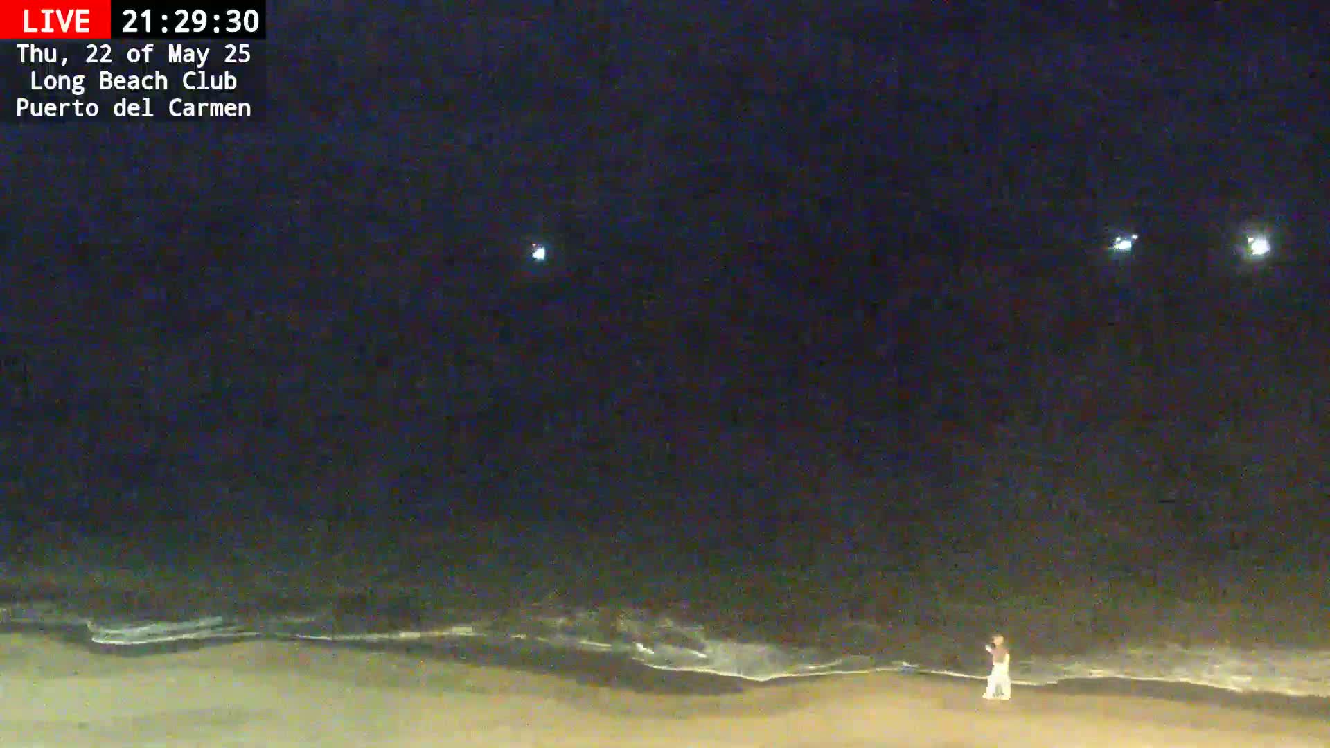 A person stands on a dark beach at night under a clear, dark sky with faint lights visible in the distance and small waves lapping at the shore.