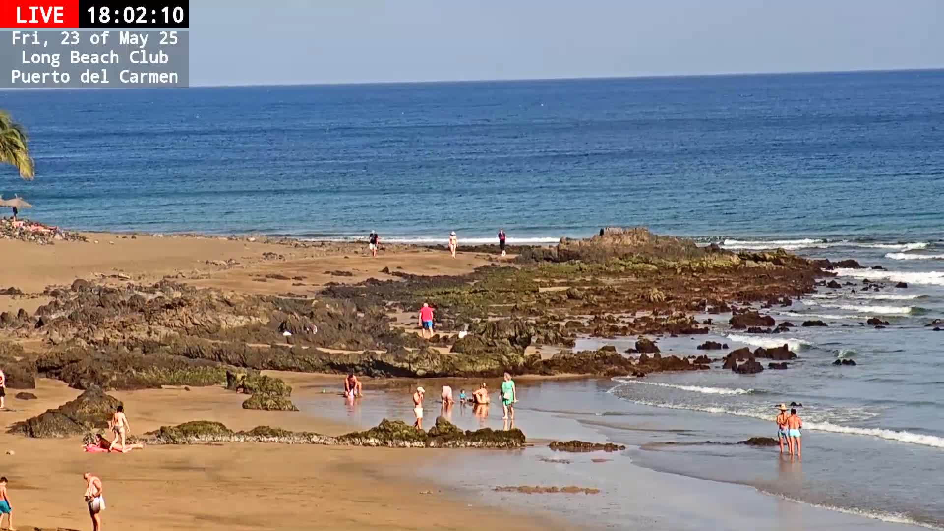 A sandy beach with dark rocks and calm ocean water under a sunny sky, with several people enjoying the shore.